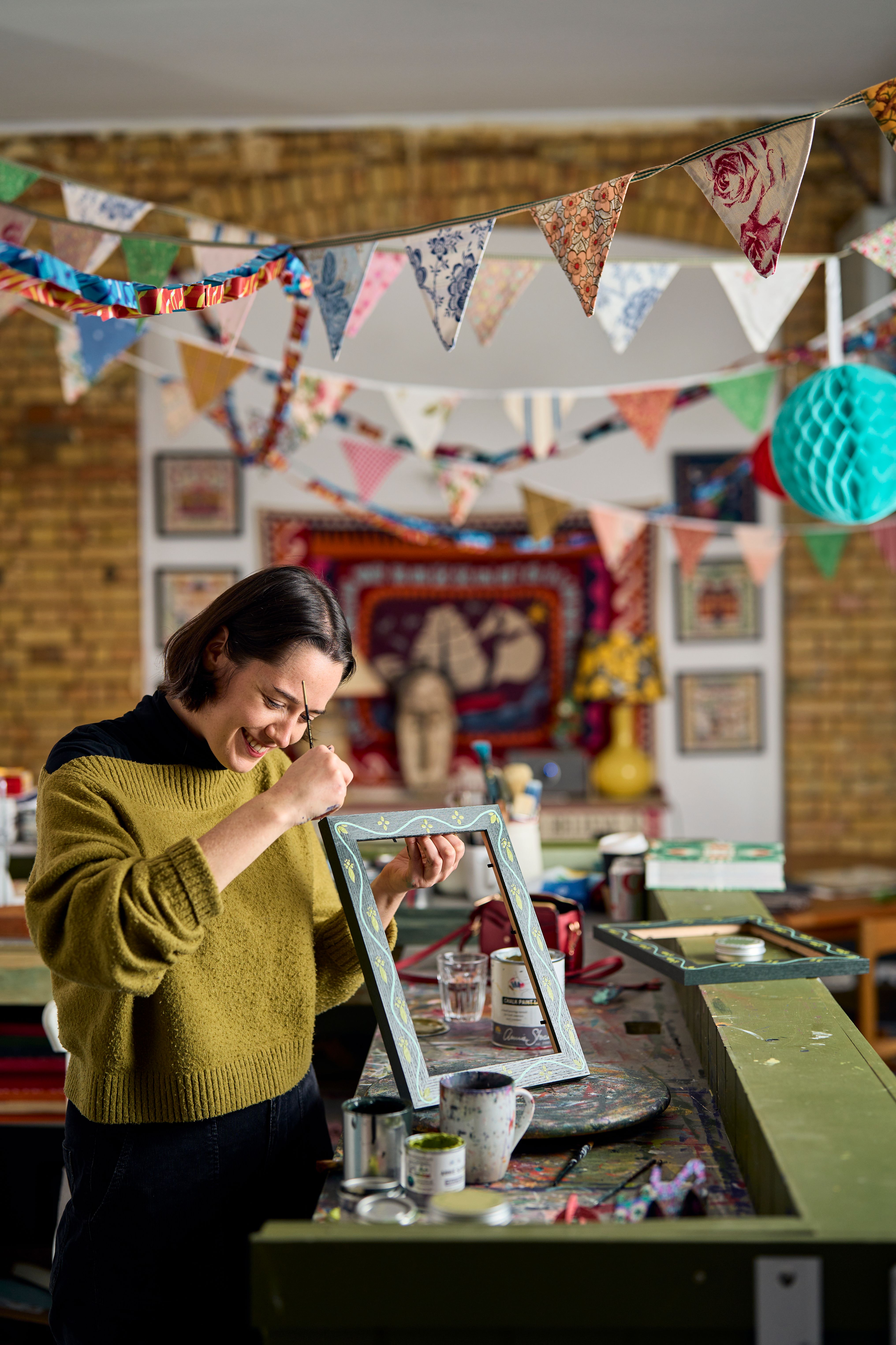 Katy in the studio at her painting station with bunting above, hand painting a frame for an embroidery kit from The Fabled Thread