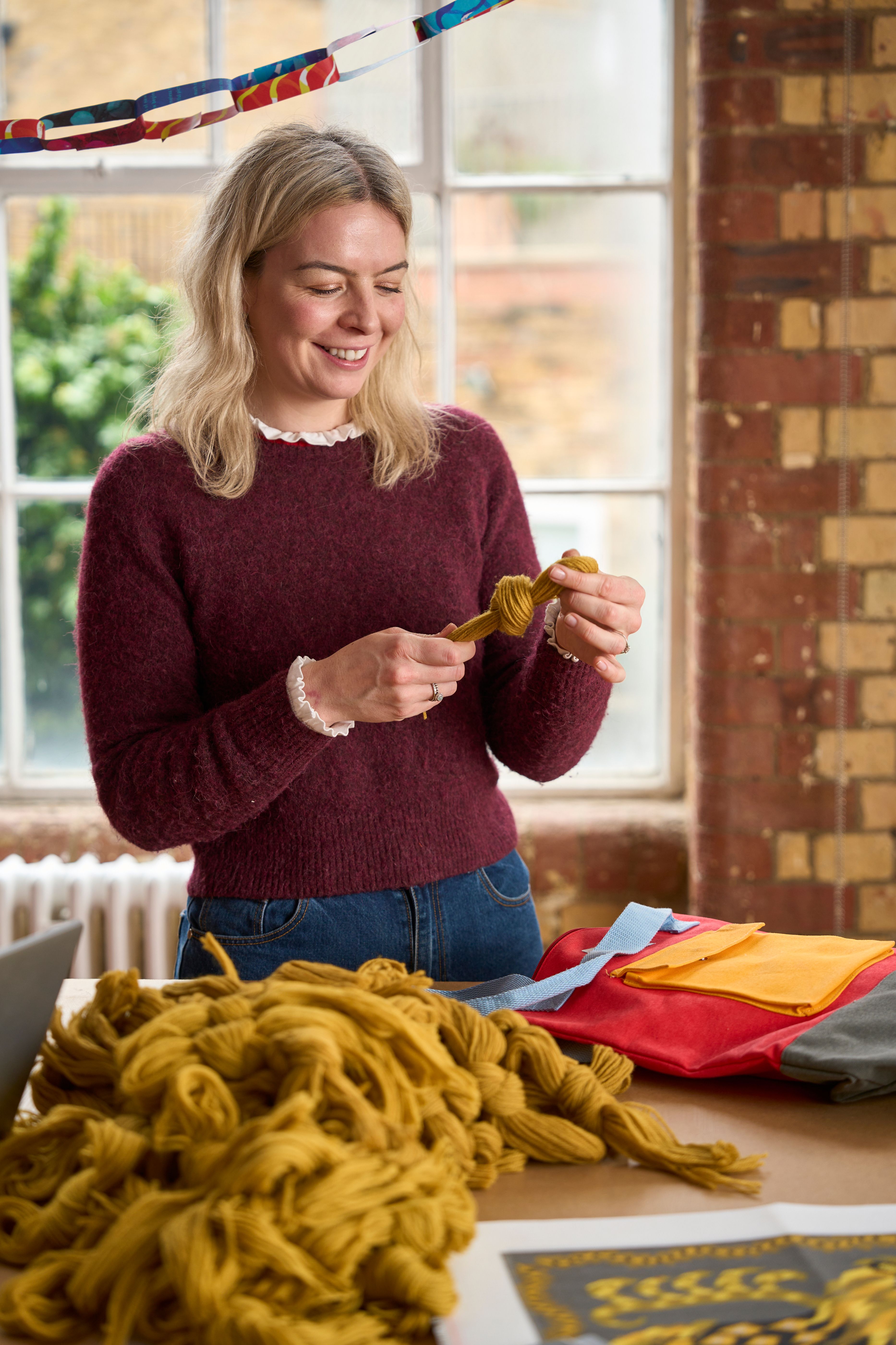 Georgina from The Fabled Thread Team in the Studio hanking mustard yellow wool for needlepoint kits with paper chains above her