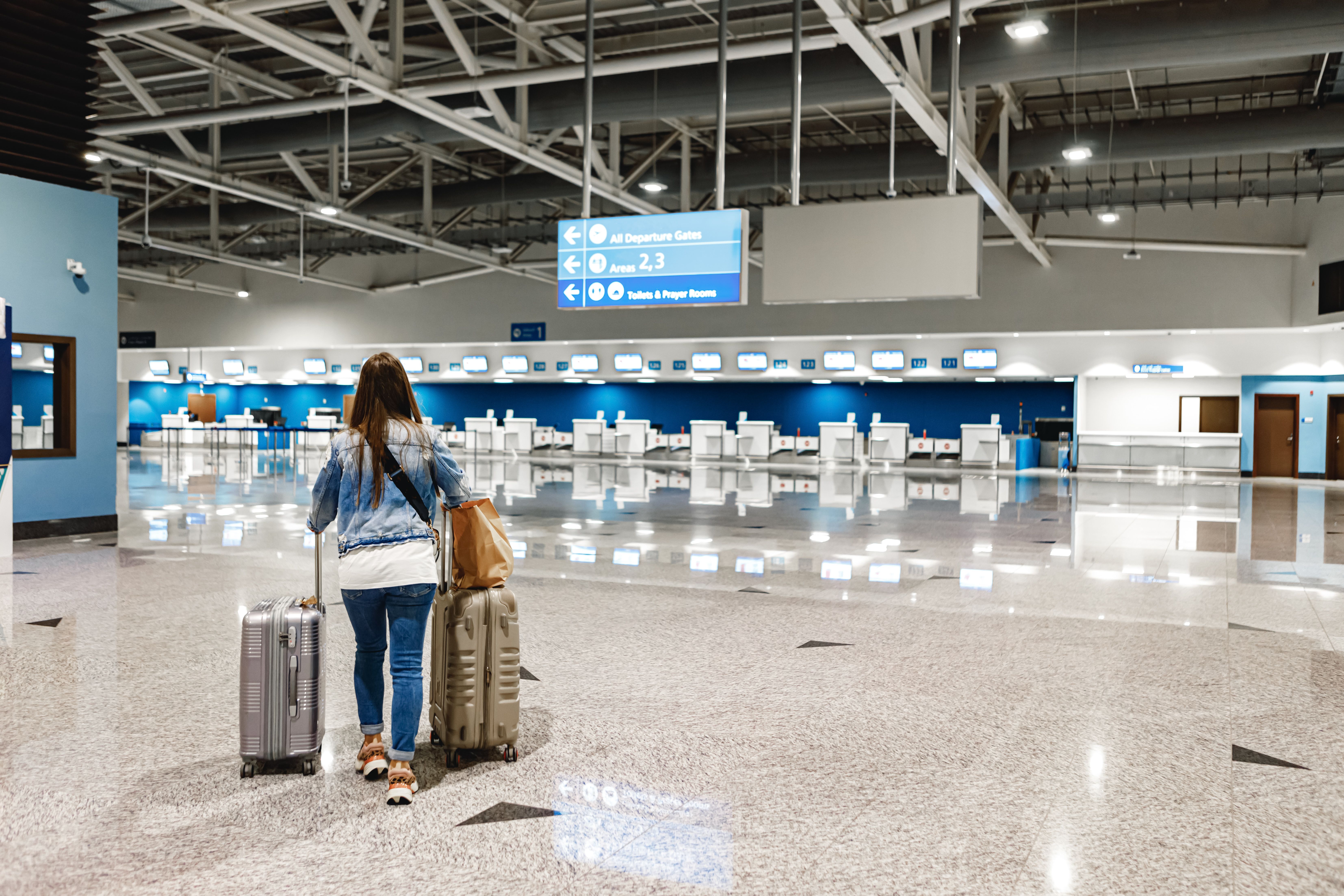 A woman pulls two suitcases through a spacious airport terminal towards blue check-in counters.