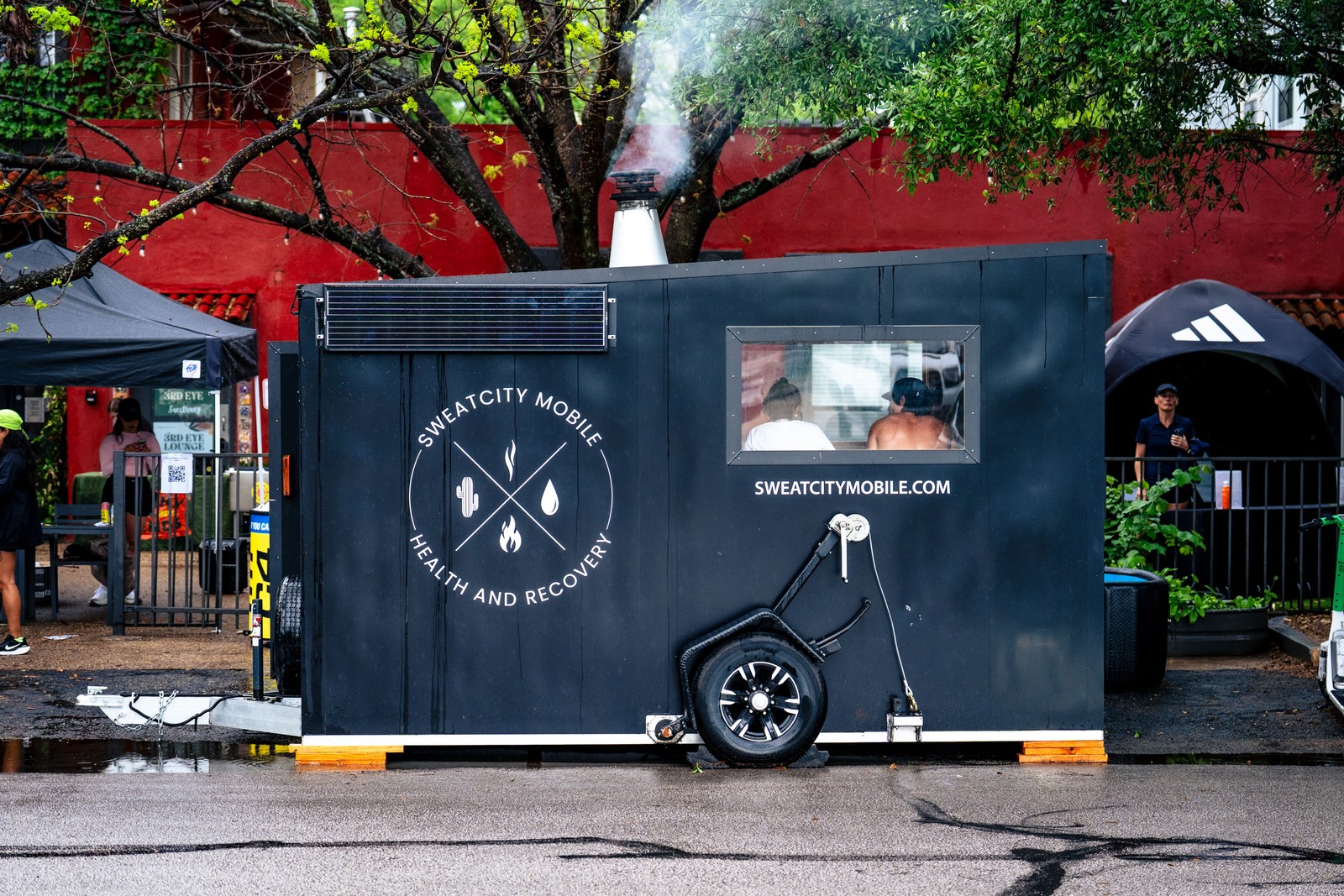 A premium mobile cedar sauna on a trailer setup alongside two sleek cold plunge tubs on an outdoor patio courtyard