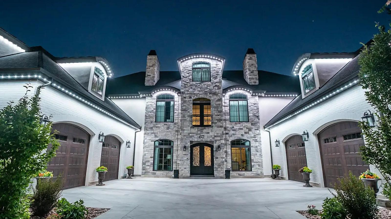 White permanent eave lights outlining a two-story white and stone home at night.