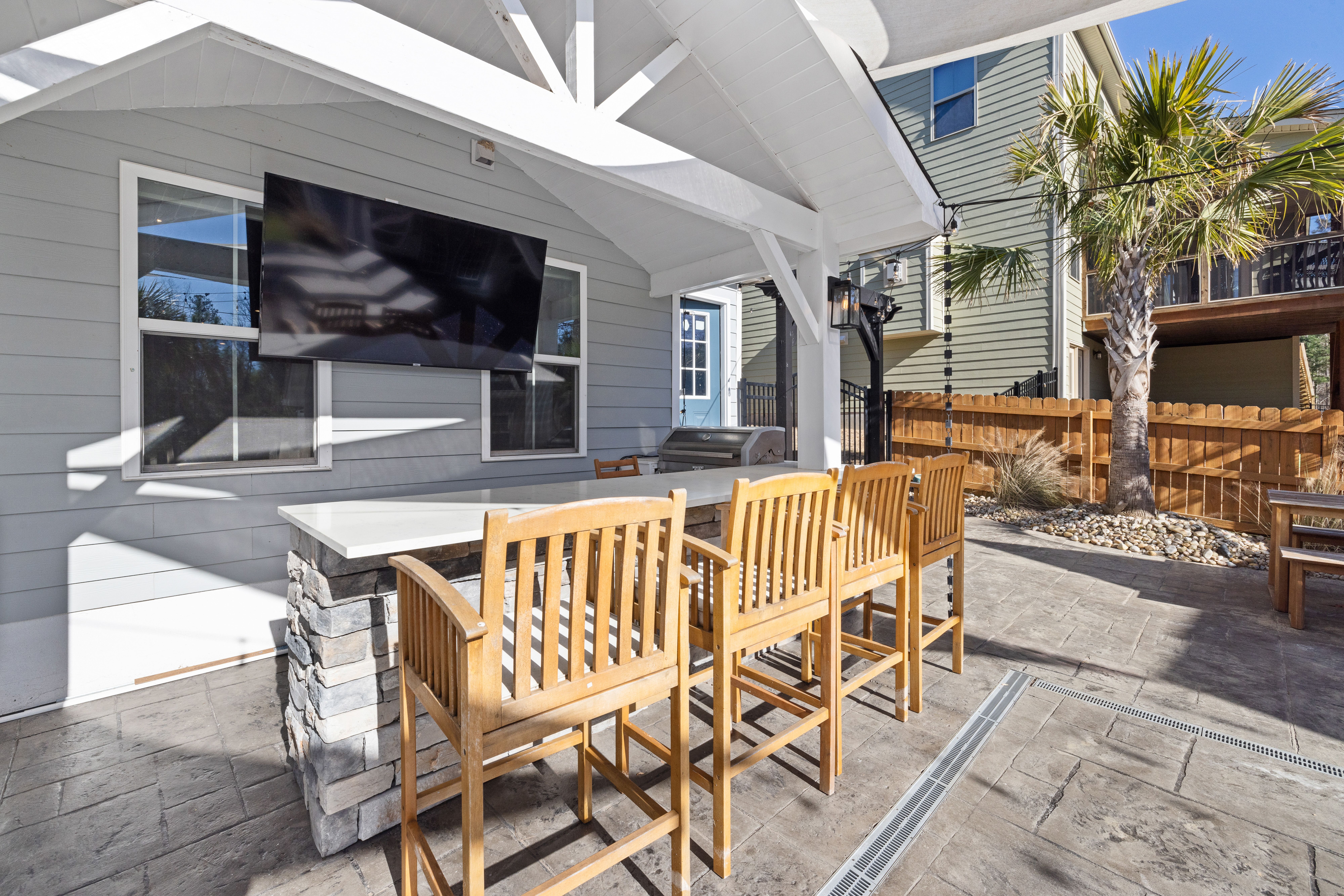 Covered residential patio with a wall-mounted television, bar seating, grill, and open backyard.