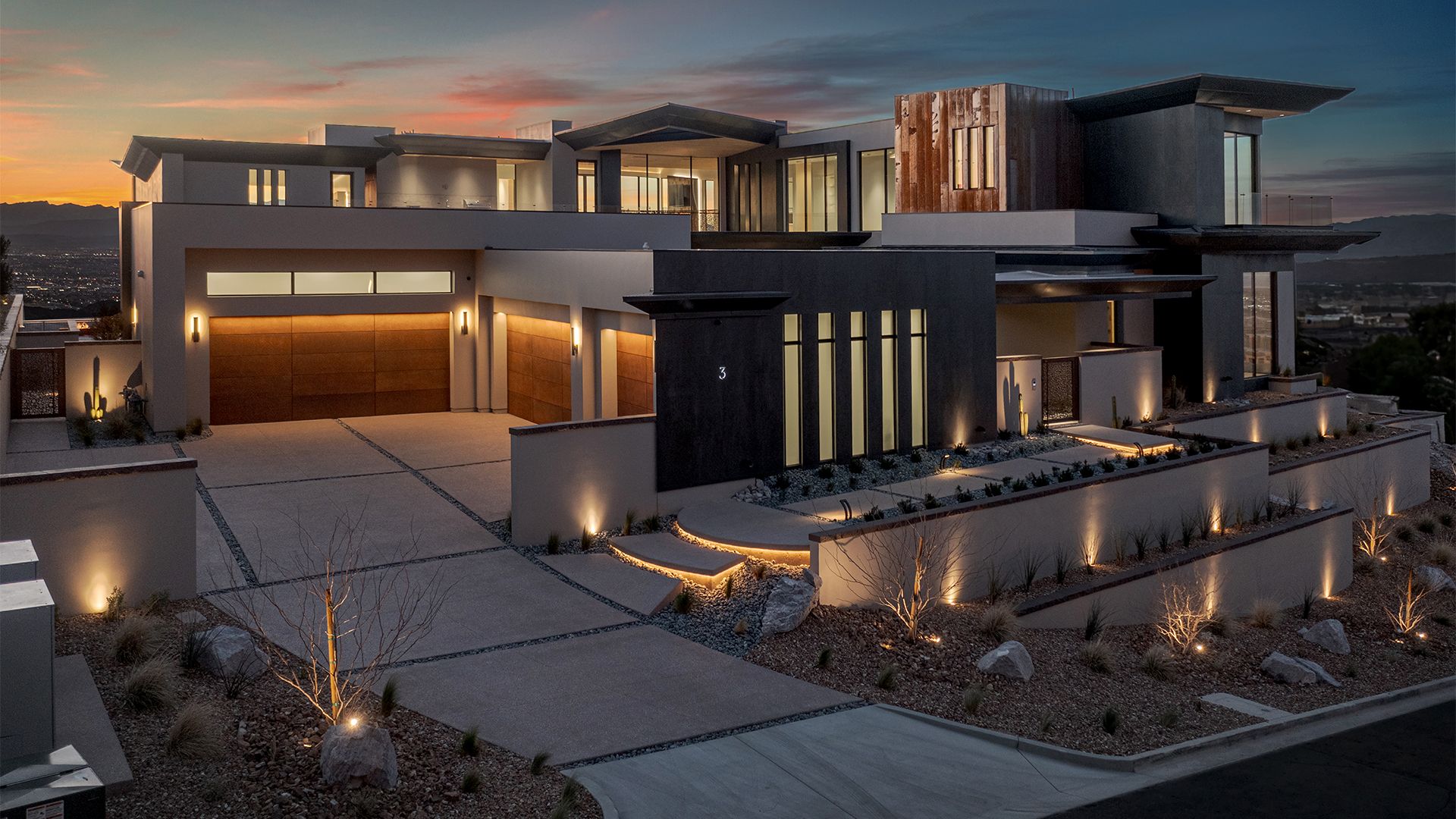 Modern custom home at dusk with warm exterior lighting along trees, walls, steps, and the driveway.