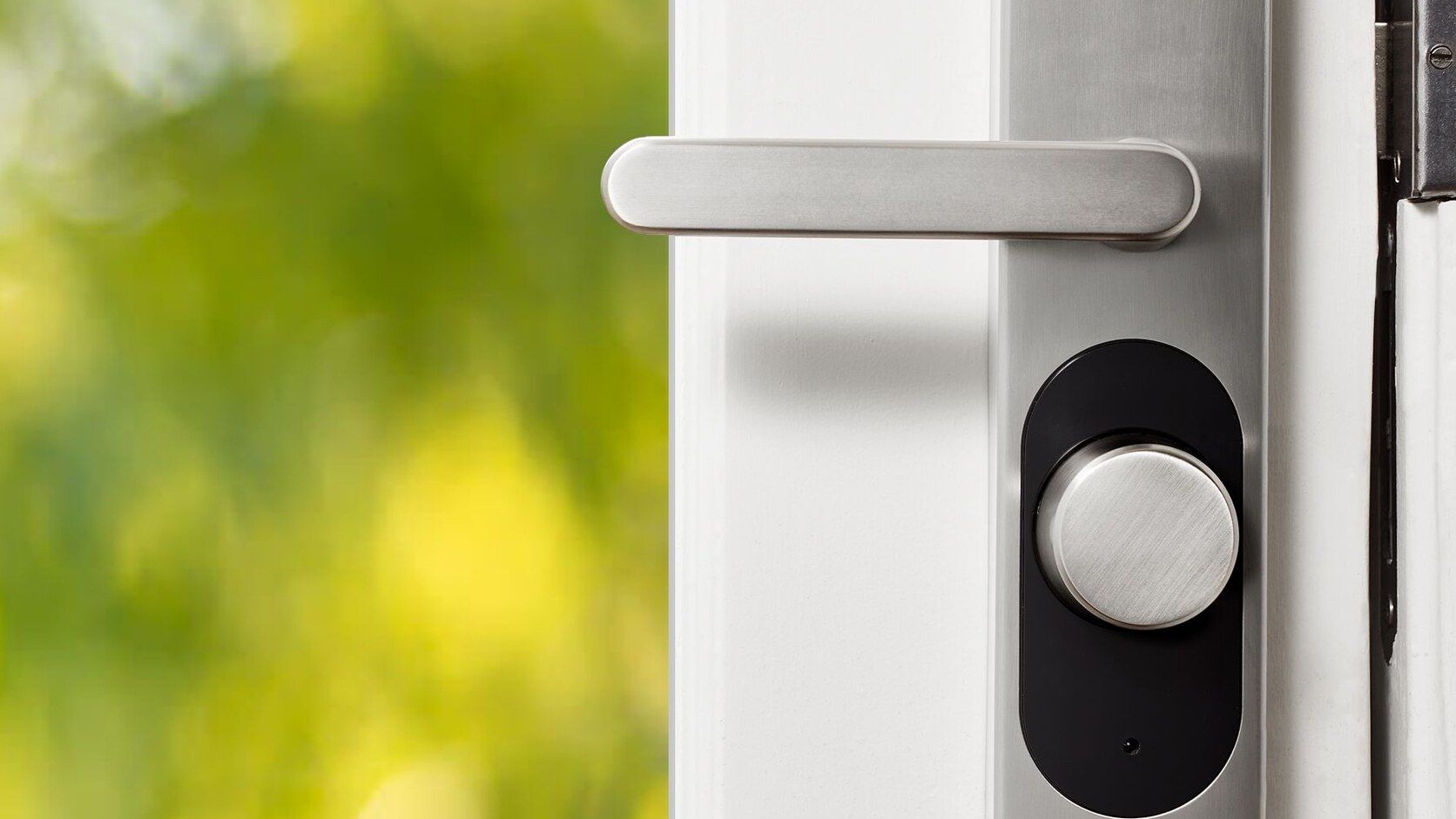 Brushed-metal smart lock hardware on a white residential door with green foliage outside.