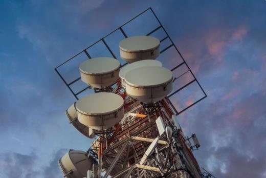 A low-angle view of a communication tower covered in multiple white dish and panel antennas against a cloudy sky.