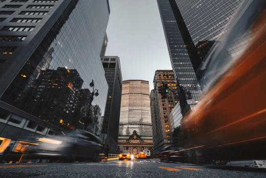 Motion-blurred cars on a city street at dusk, with towering skyscrapers and Grand Central Terminal in the background.