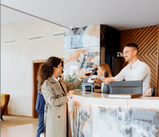 A woman uses her phone for contactless payment at a reception desk, assisted by a smiling male staff member.