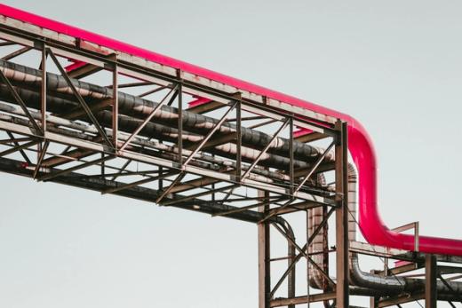 Bright pink industrial pipe on a metal truss with smaller pipes against a light sky.