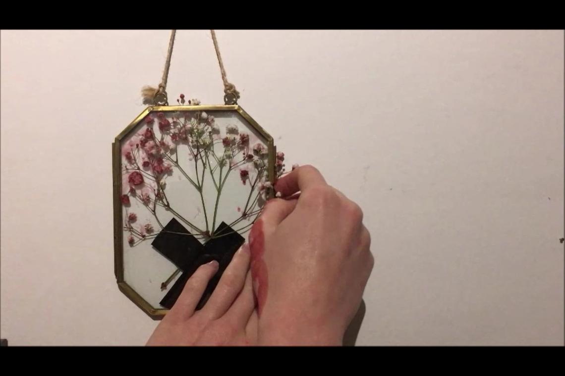 A film still from “[this is not] a piece about my disability”. A glass picture frame with brass edges on a white wall.  Blood soaked flowers have been roughly gaffer-taped into the frame. A pair of blood stained hands are latching the frame shut.