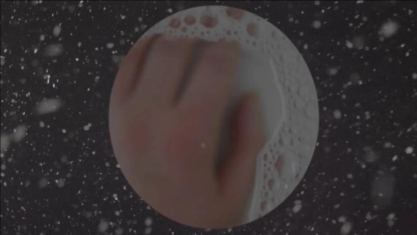 Still from the video. Dark background with white particles. Circle cutout image in centre showing a hand and water bubbles.