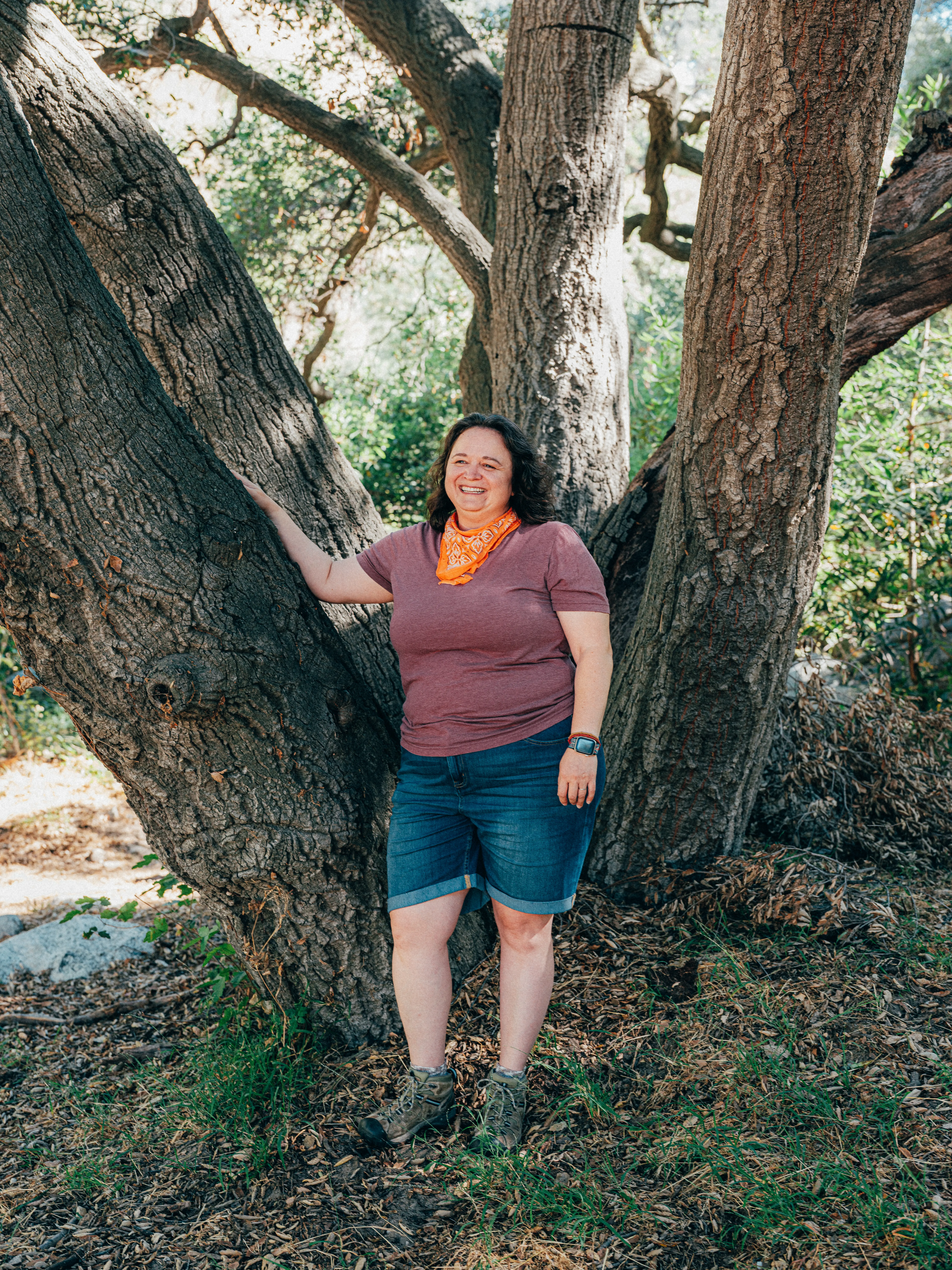 A woman stands beside a large tree, resting her hand on its trunk