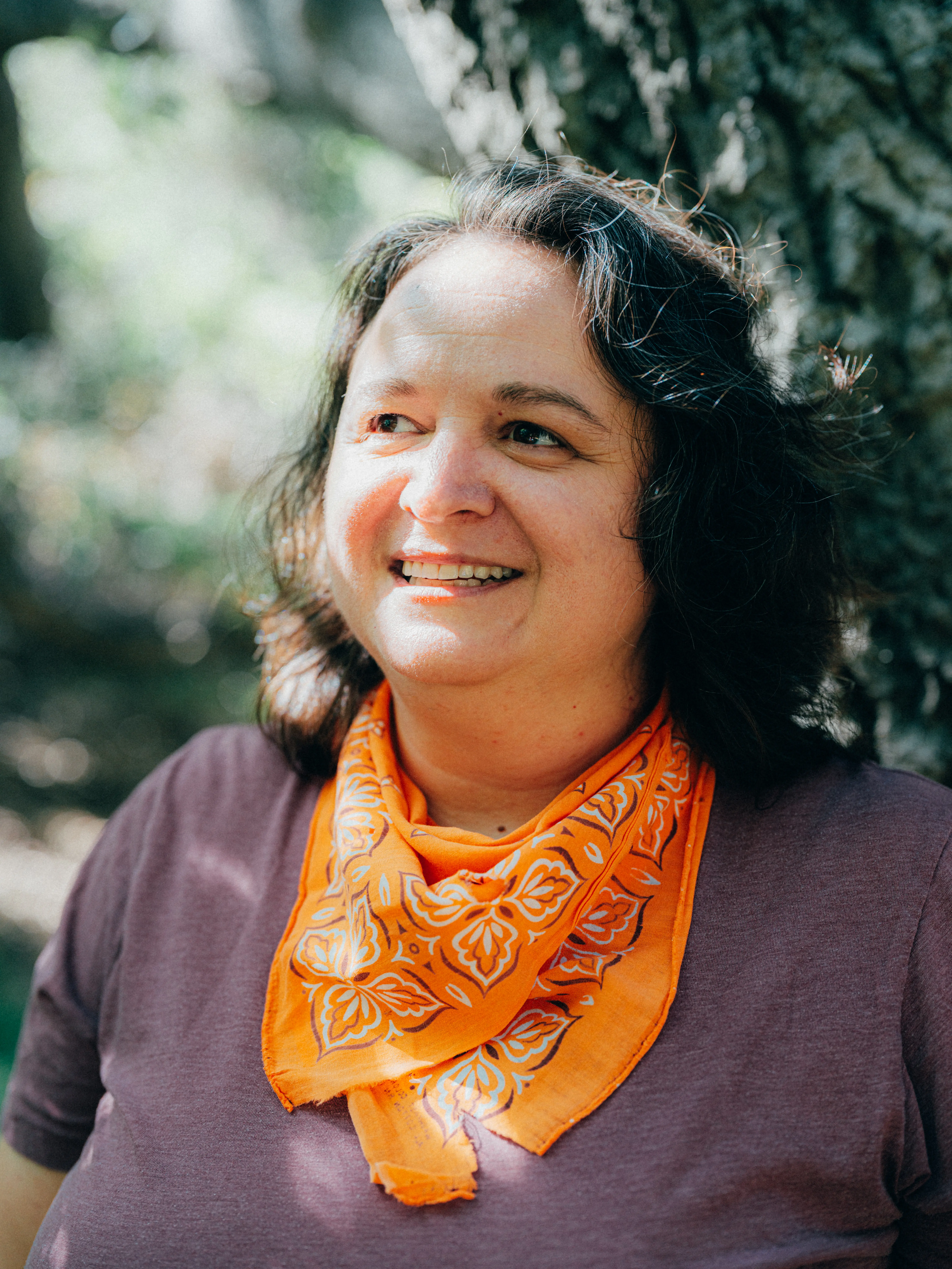 A woman stand against a large tree trunk, wearing a maroon shirt and orange bandana tied around her neck 