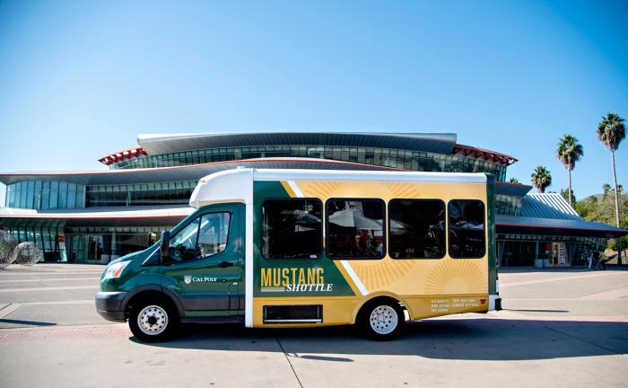 A green and gold bus with the words Mustang Shuttle on its side in front of the Performing Arts Center