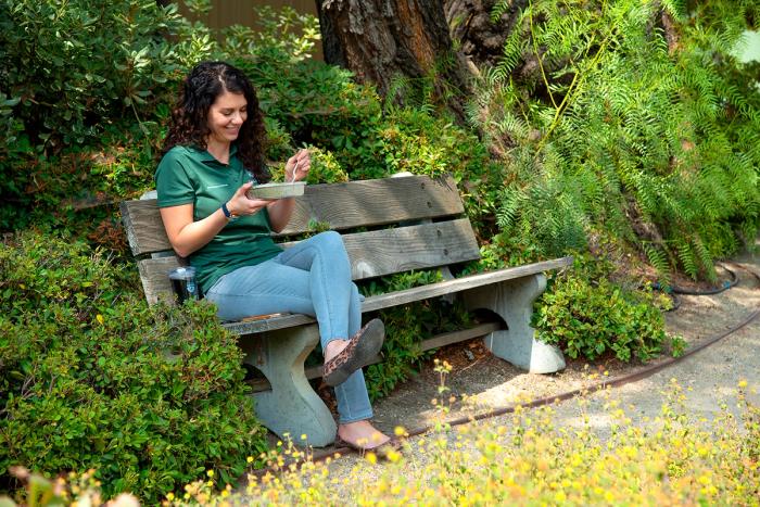 A campus staff member is sitting on a bench in front of some trees eating lunch.
