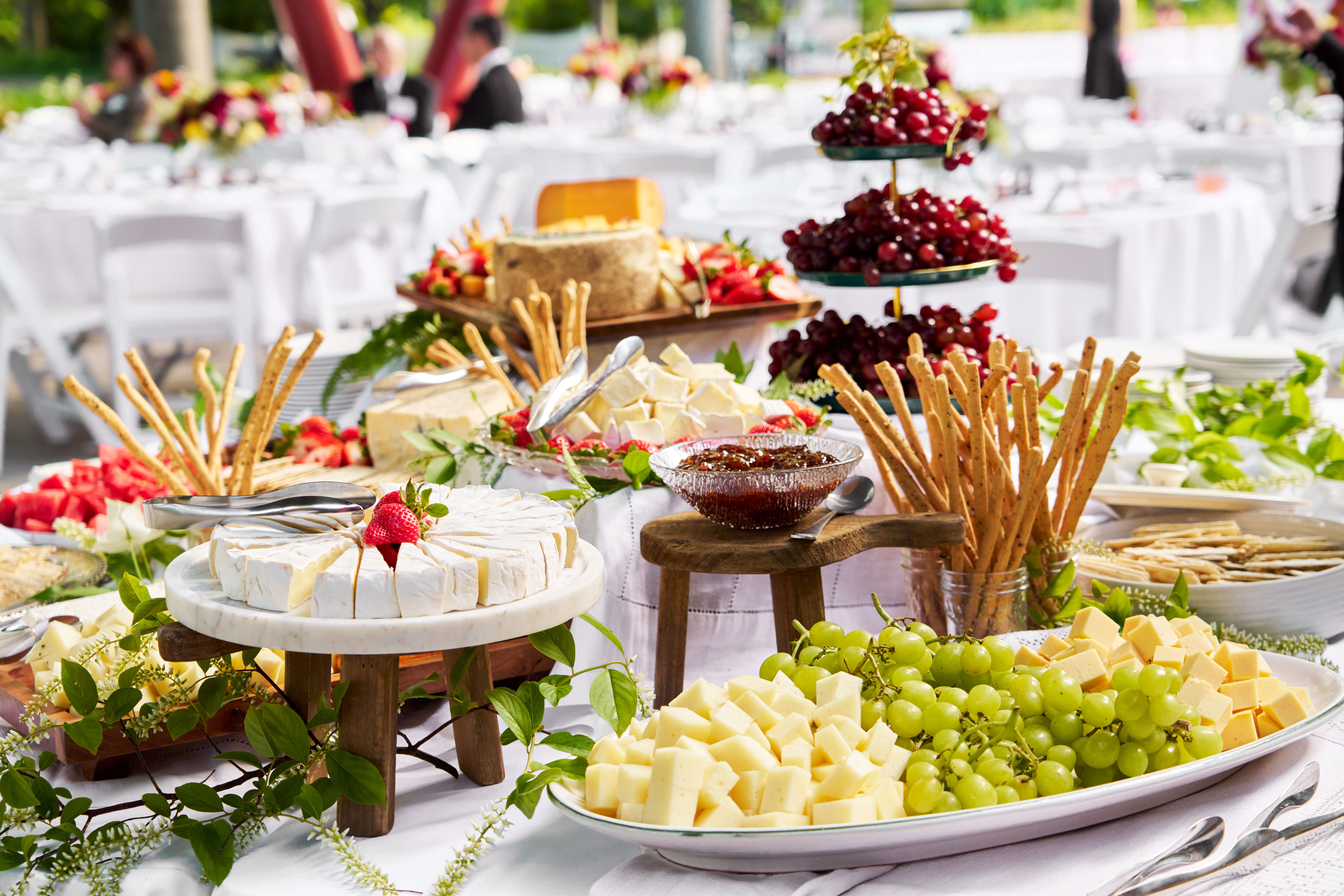 An outdoor catering table laden with cheeses, grapes, fruit, and breadsticks.