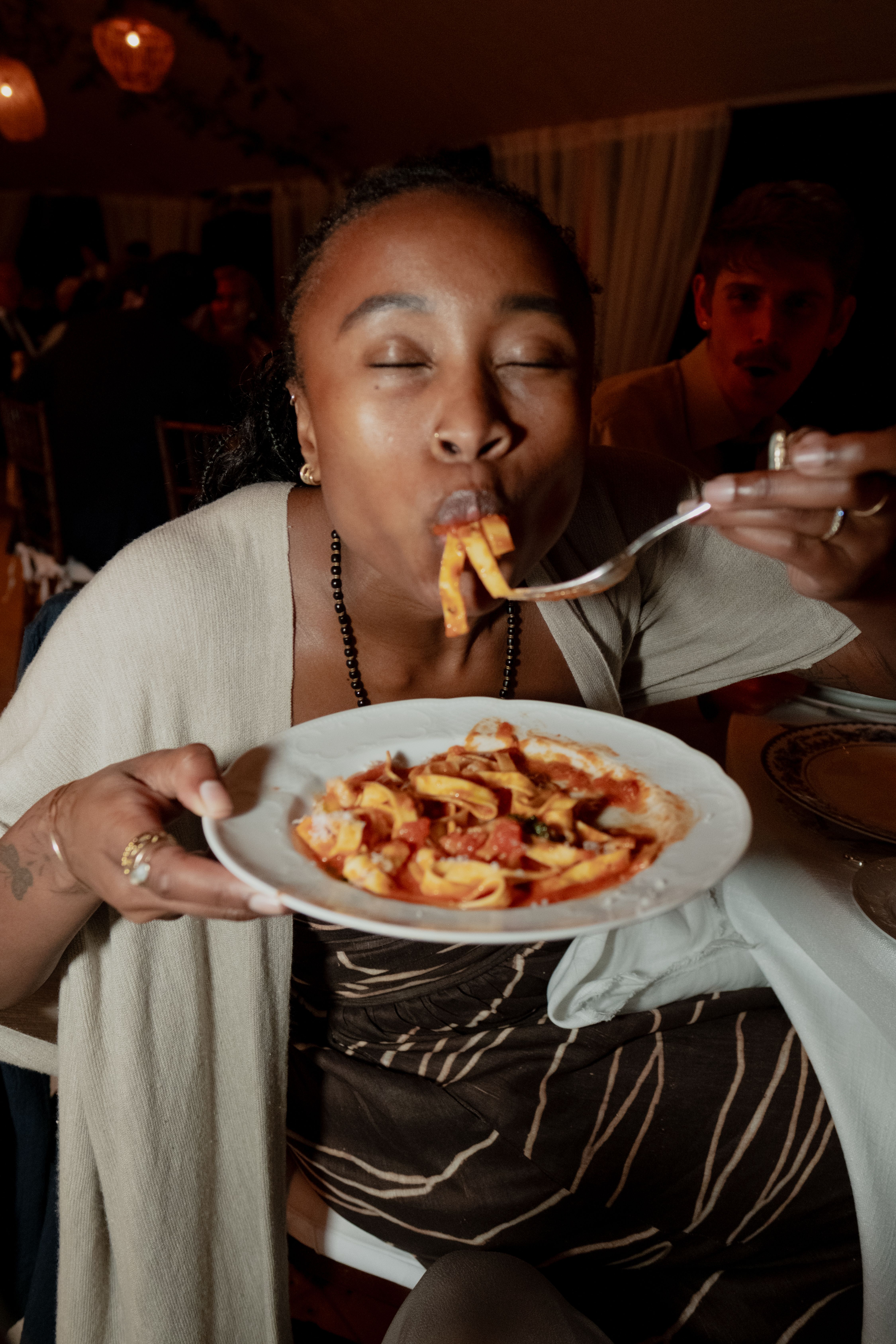 A person with closed eyes eagerly eats pasta from a fork, holding a plate, with a surprised man in the background.