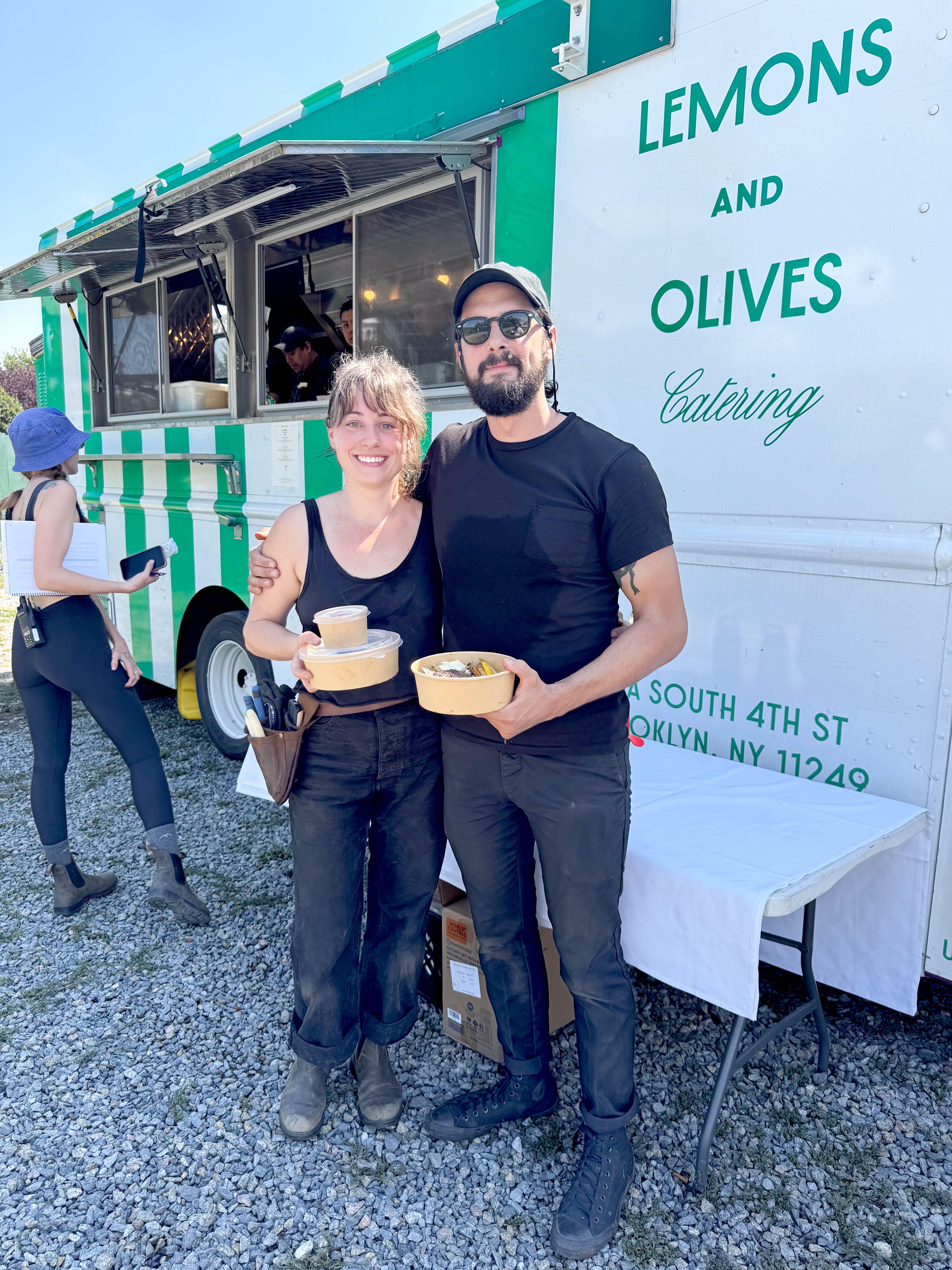 A smiling man and woman hold food containers in front of a white food truck with "Lemons and Olives Catering" written on its side.