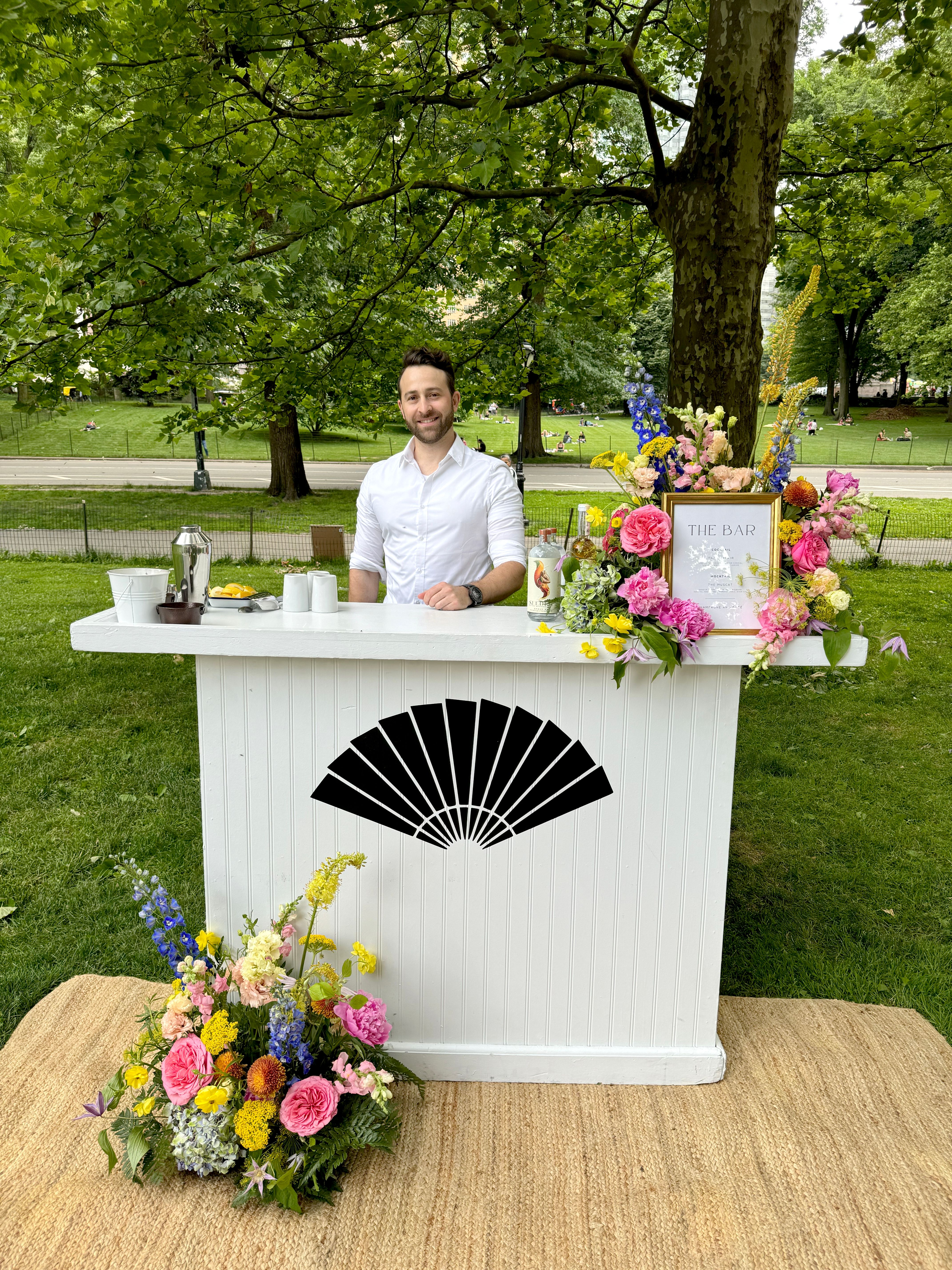 Mobile cocktail bar with floral decor set up for an outdoor catered event in a park.