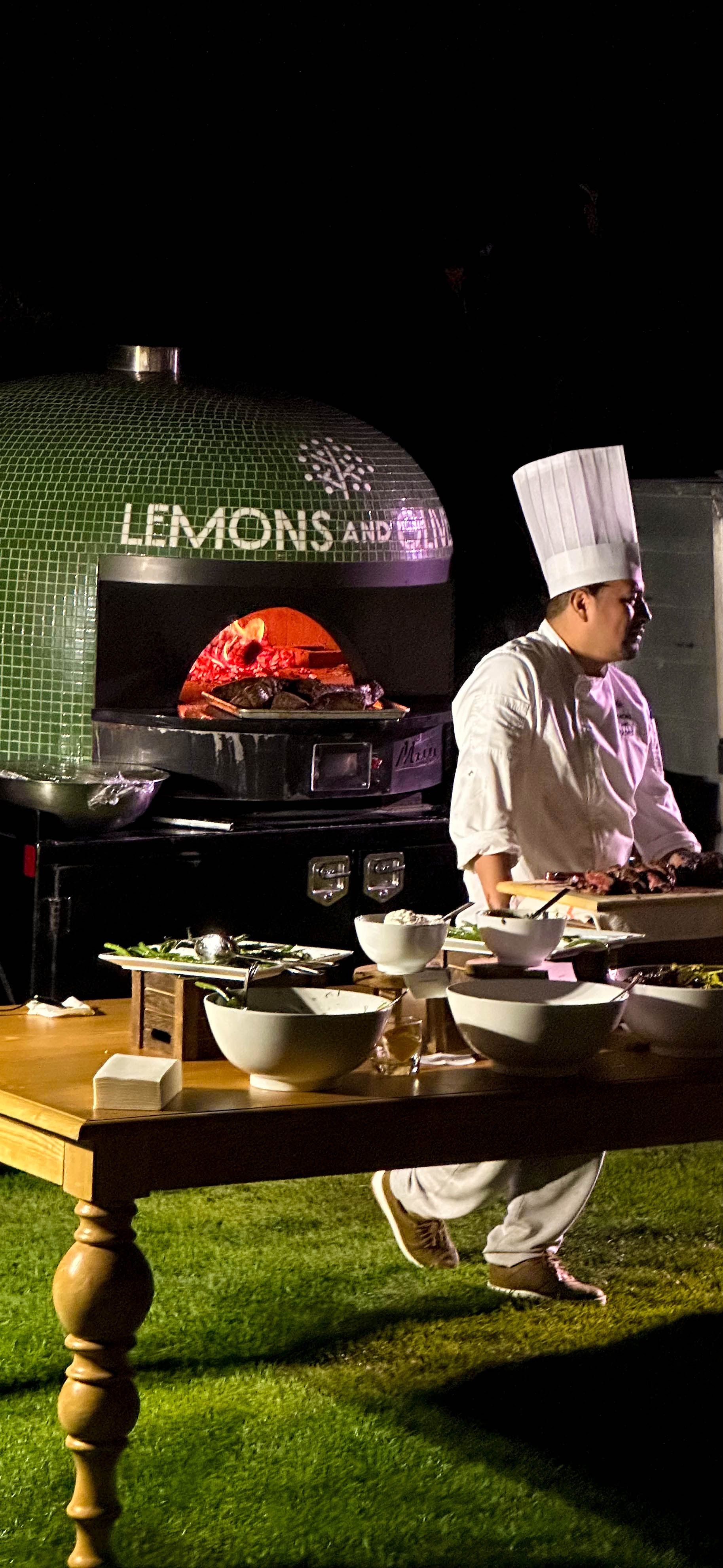 Chef preparing dishes beside a wood-fired pizza oven at a live catering station during an event.