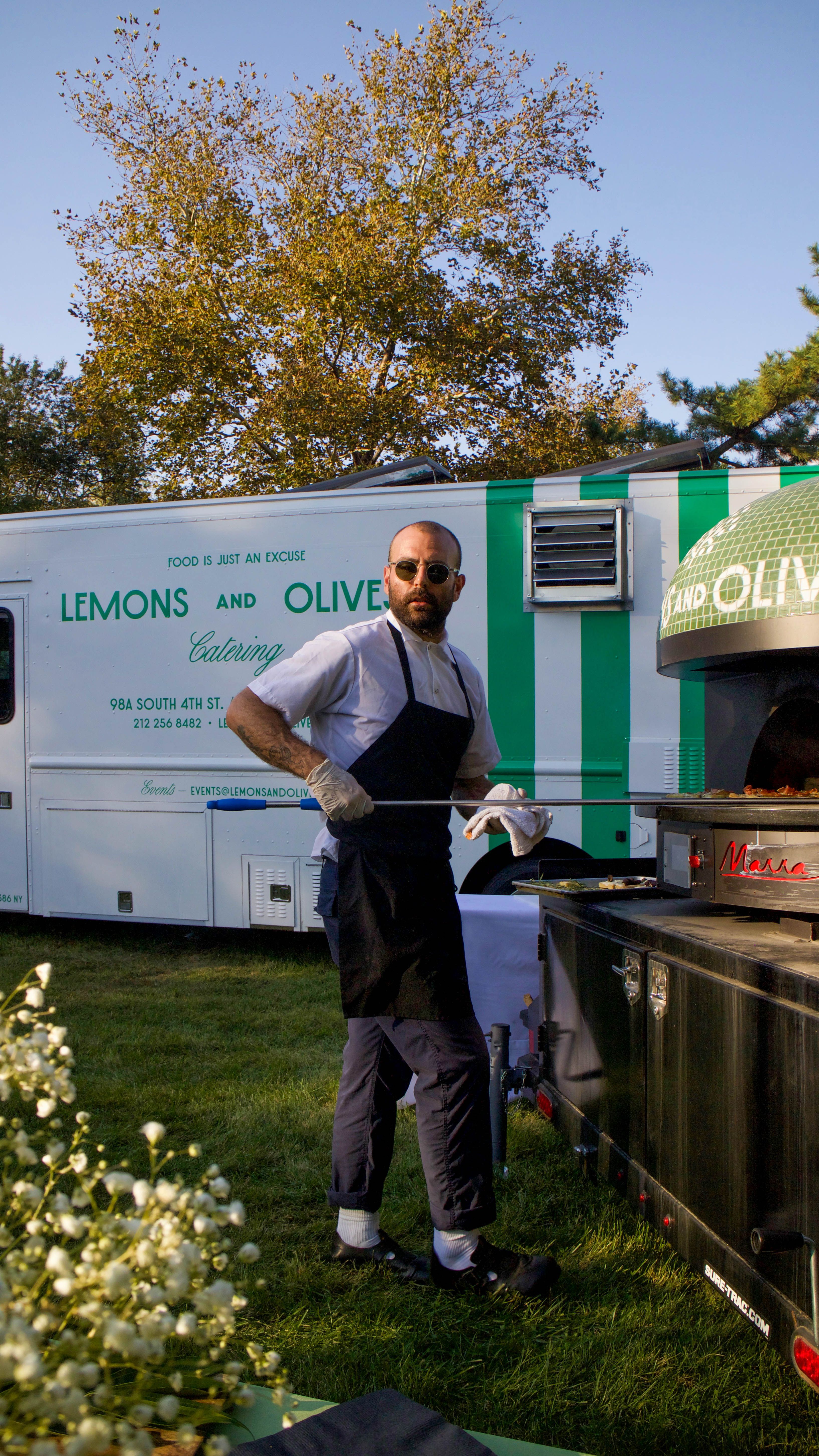 A chef with sunglasses and an apron operates a wood-fired oven next to a 'Lemons and Olives' catering food truck on a grassy lawn in the Hamptons.