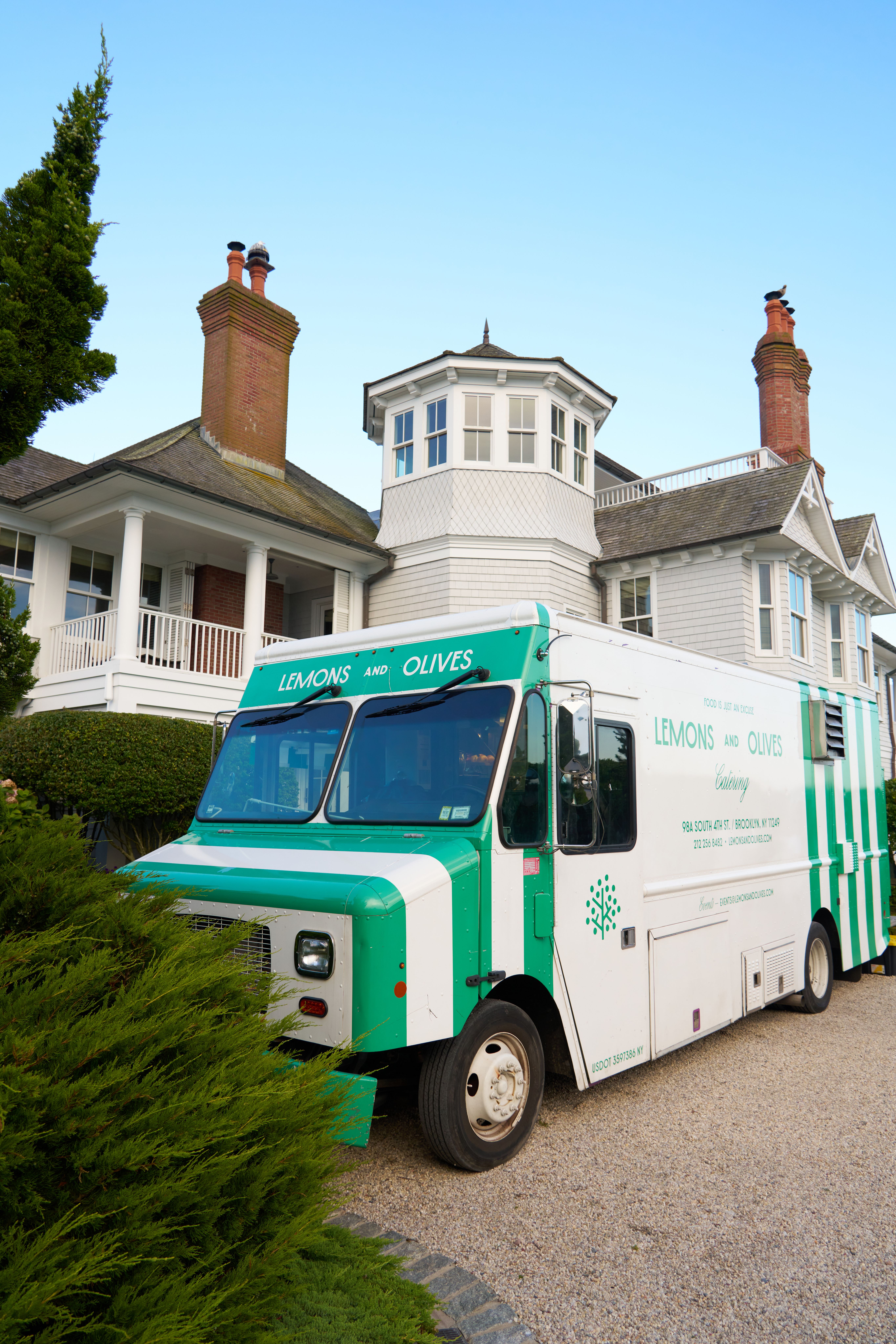 A white and green striped "Lemons and Olives" catering food truck parked on a gravel driveway in front of a large house.