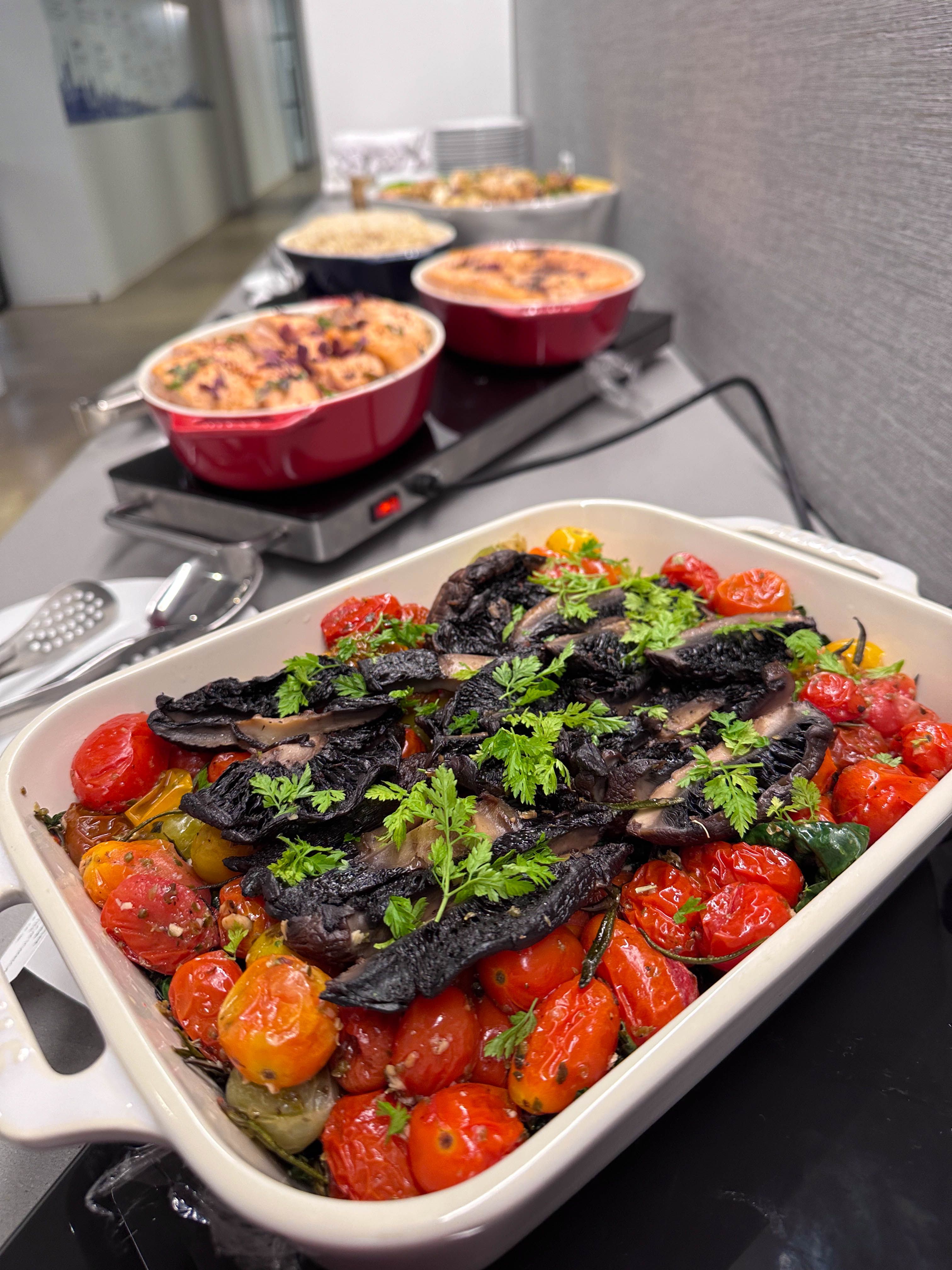 Catering tray of roasted mushrooms and cherry tomatoes garnished with herbs at a buffet station.