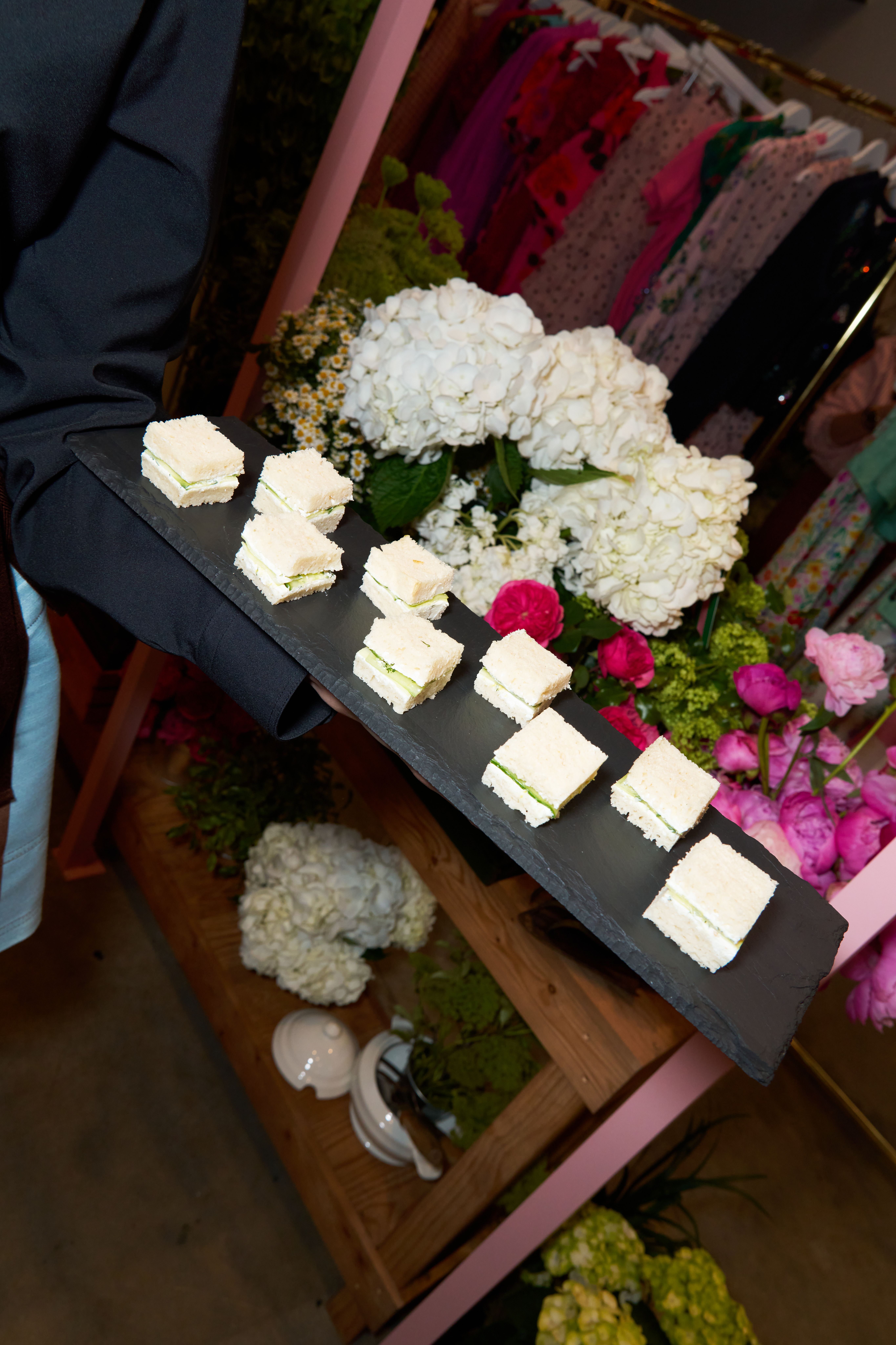 A server holds a black slate tray with cucumber sandwiches, with floral decorations and clothing racks in the background.