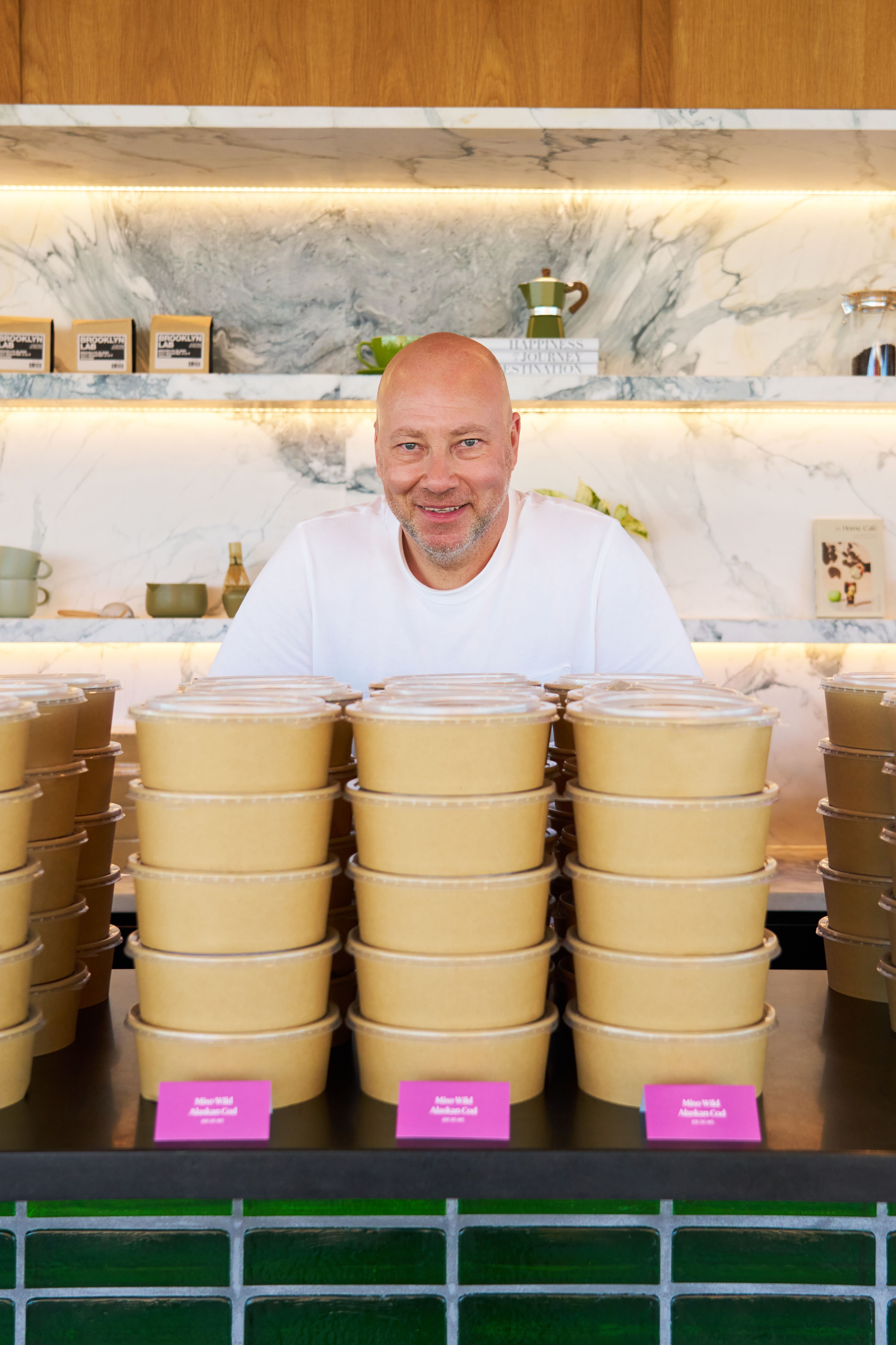 A smiling bald man in a white t-shirt stands behind a counter stacked with takeaway containers labeled "Miso Wild Alaskan Cod."