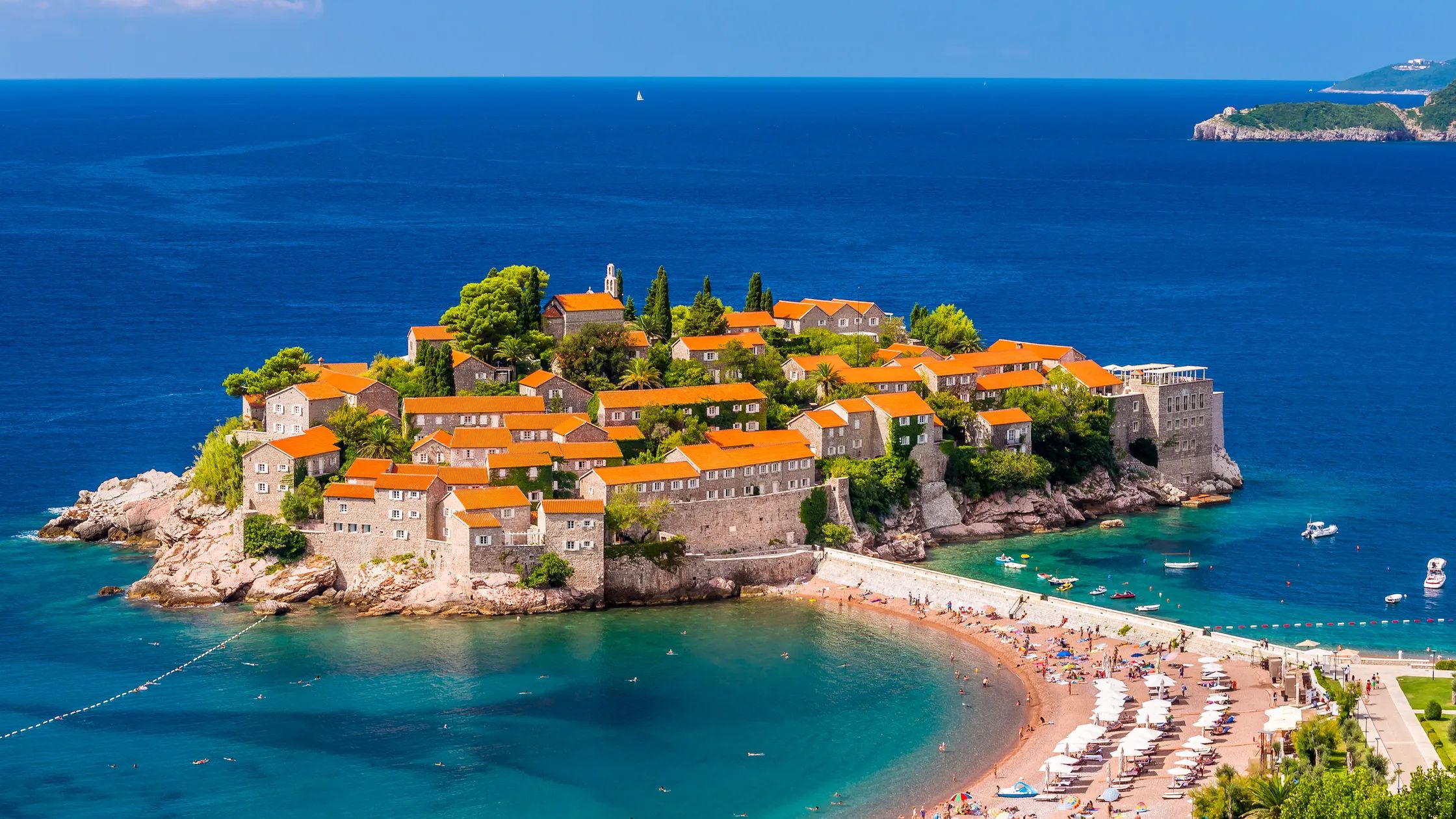 Island village with orange-roofed stone buildings, surrounded by clear blue sea, with a beach and a connecting causeway.