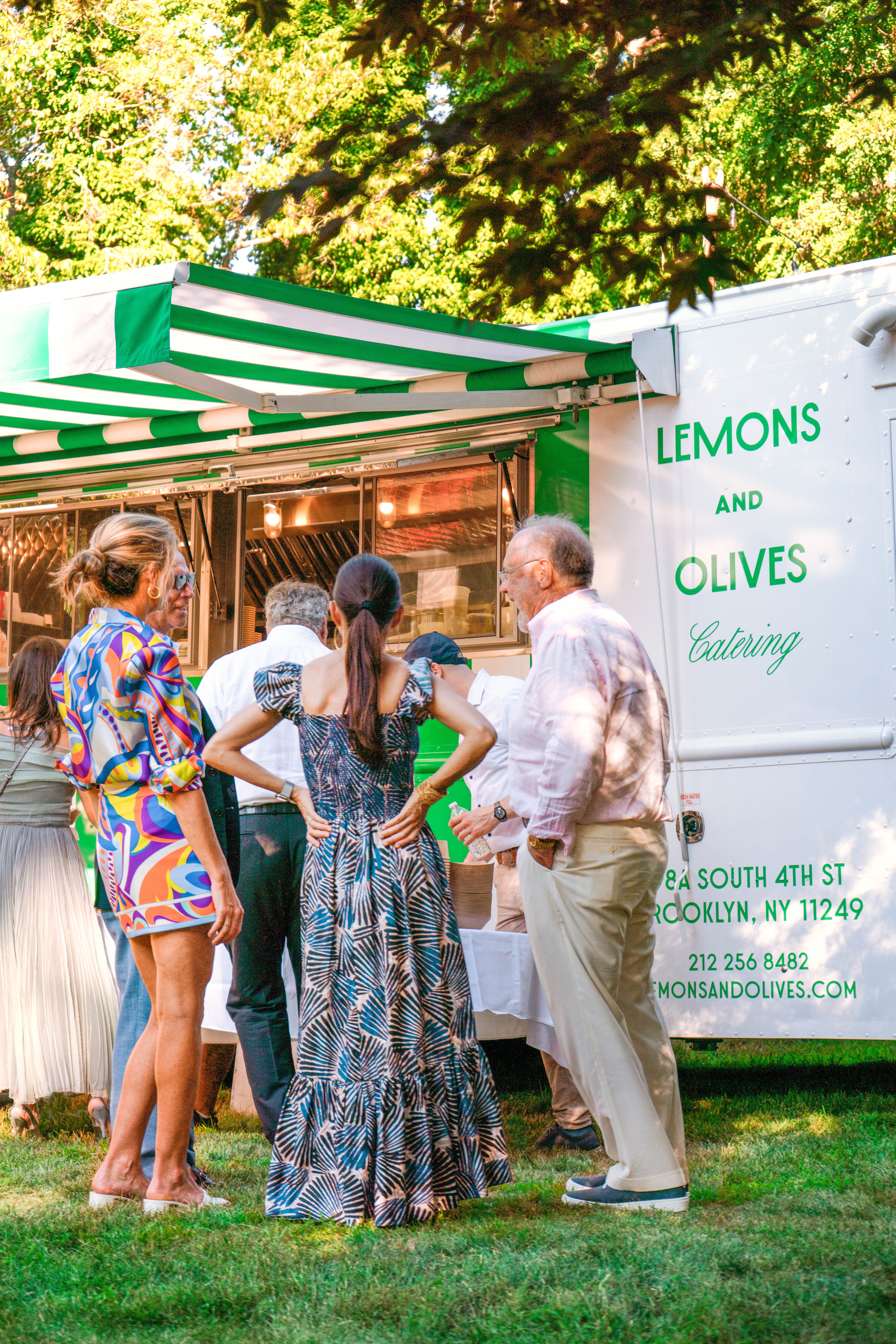 People gathered around a white food truck with a green striped awning, labeled "Lemons and Olives Catering," at an outdoor event.