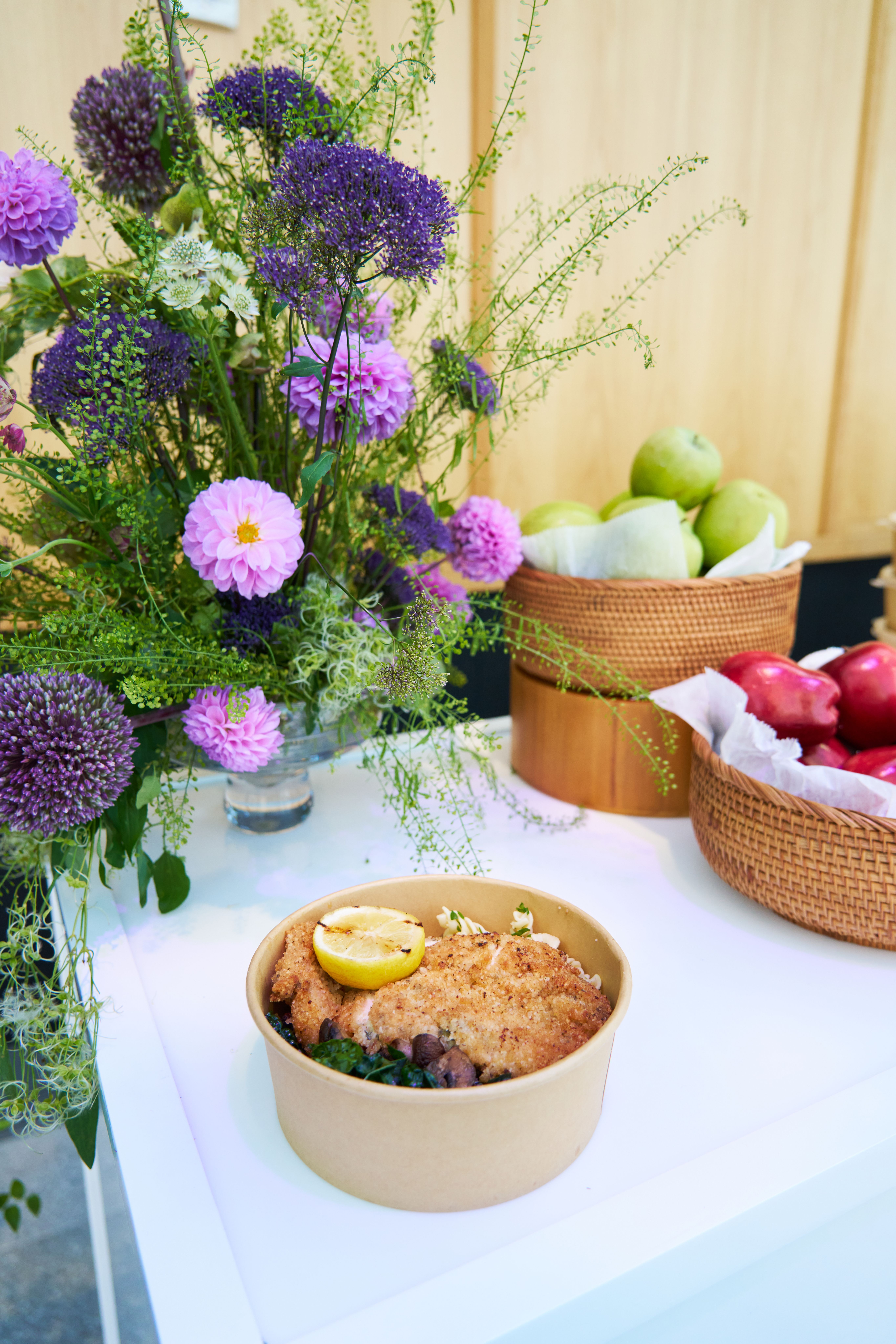 A takeout bowl with breaded chicken and a lemon slice on a table, with a purple floral arrangement and baskets of apples.
