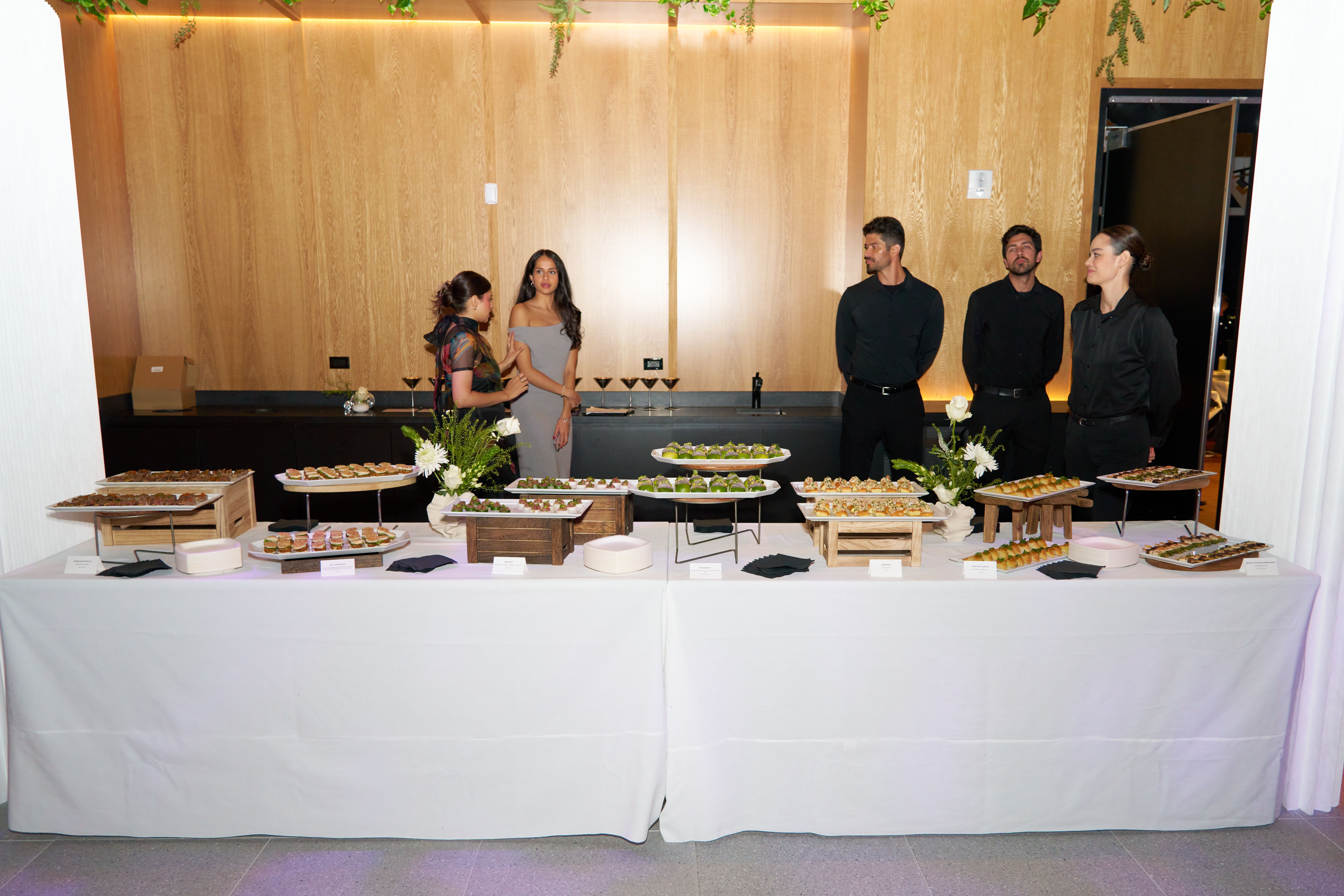 A long white buffet table displaying various appetizers, with two women and three servers behind it.