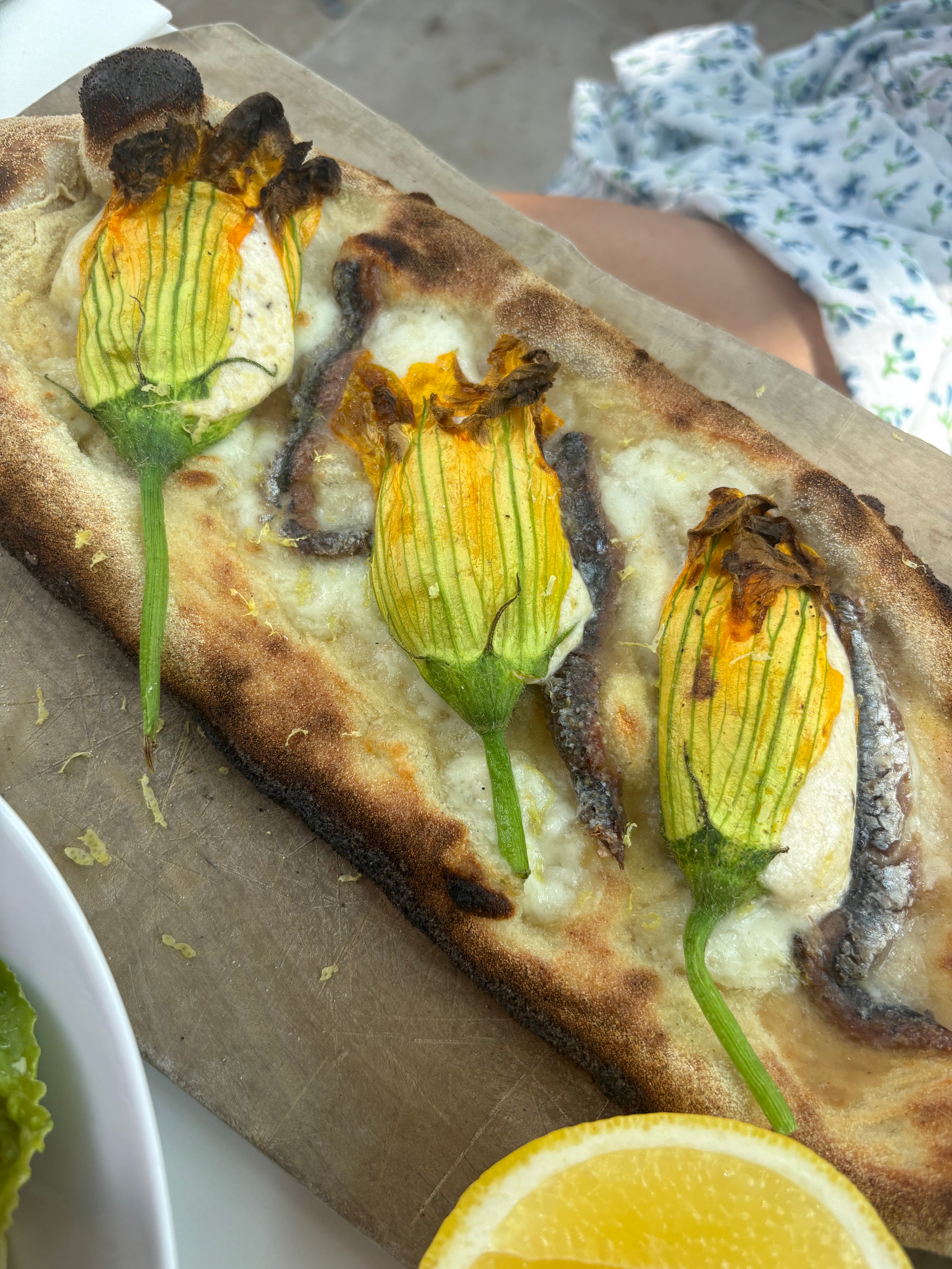 Rectangular pizza served at a wedding topped with three stuffed squash blossoms, anchovies, and cheese, on a wooden board.