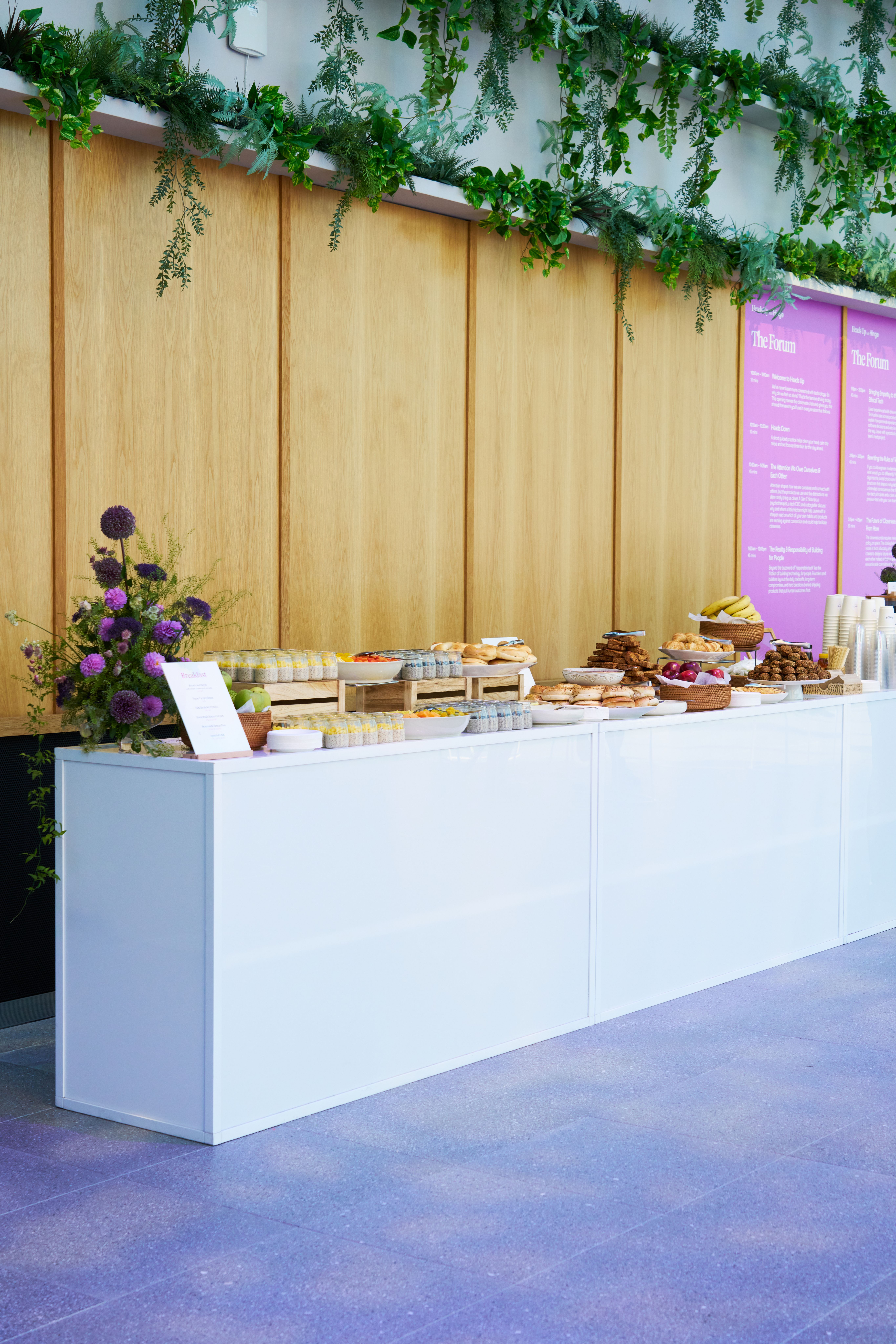 Multiple stacks of light brown takeout containers with pink labels are arranged on a counter alongside a vibrant purple floral display.