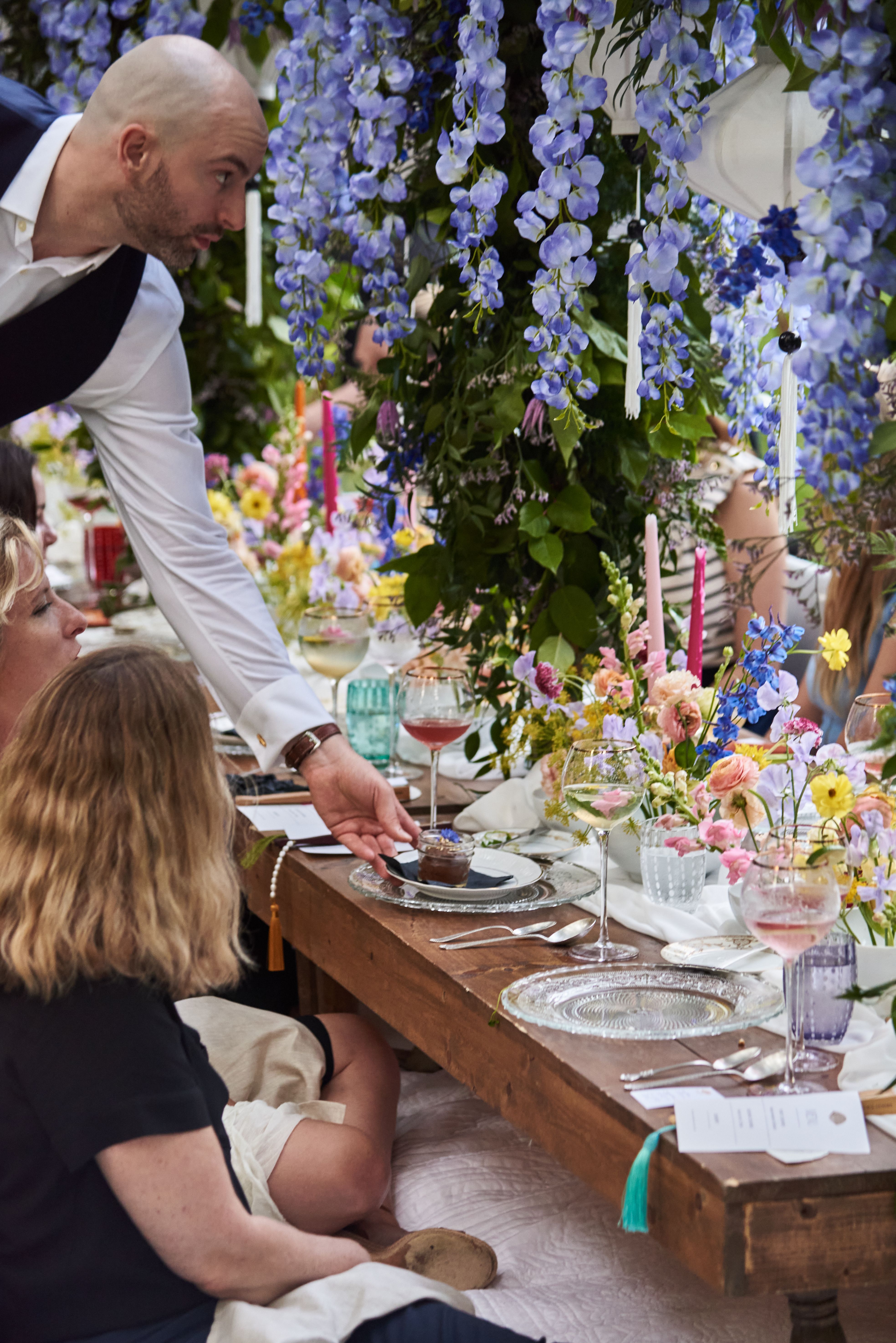 Server presenting plated mini dessert to guests at an outdoor catering dinner with floral décor.