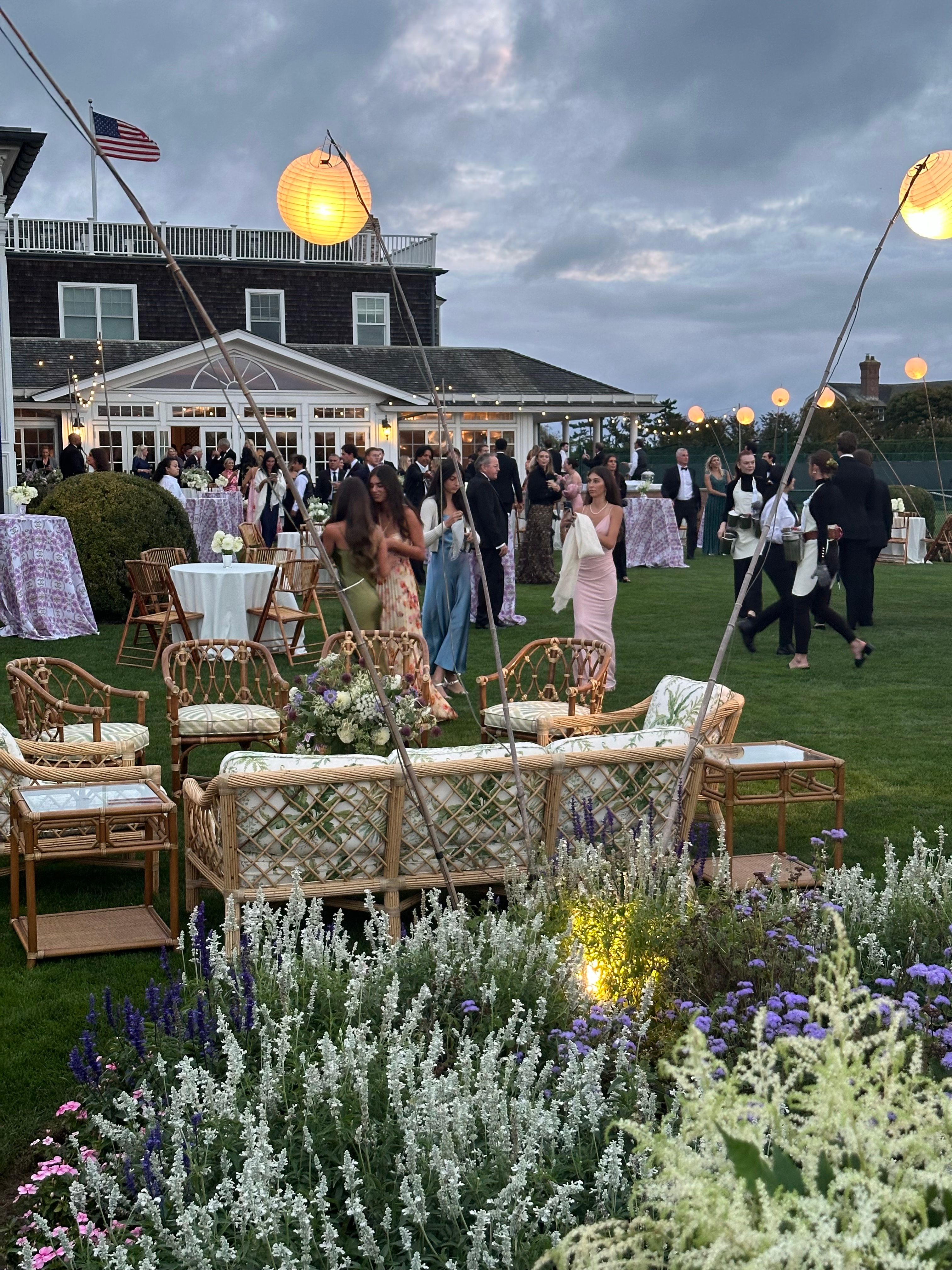 An elegant outdoor evening party at dusk, with guests mingling on a lush lawn in front of a grand building, decorated with paper lanterns, string lights, and rattan lounge furniture, with flowering plants in the foreground.