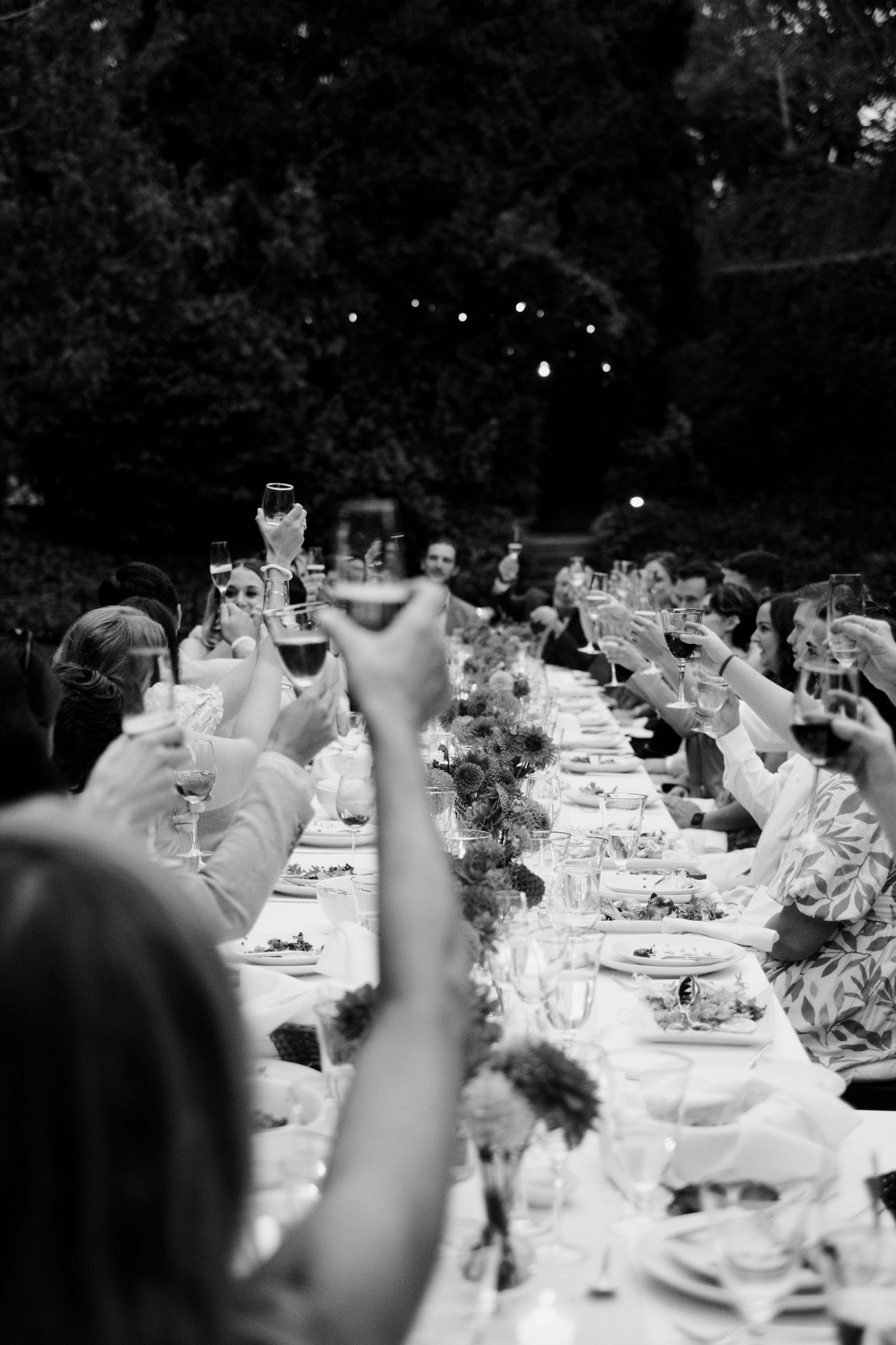 Black and white photo of guests raising glasses in a toast at a long outdoor dinner table.