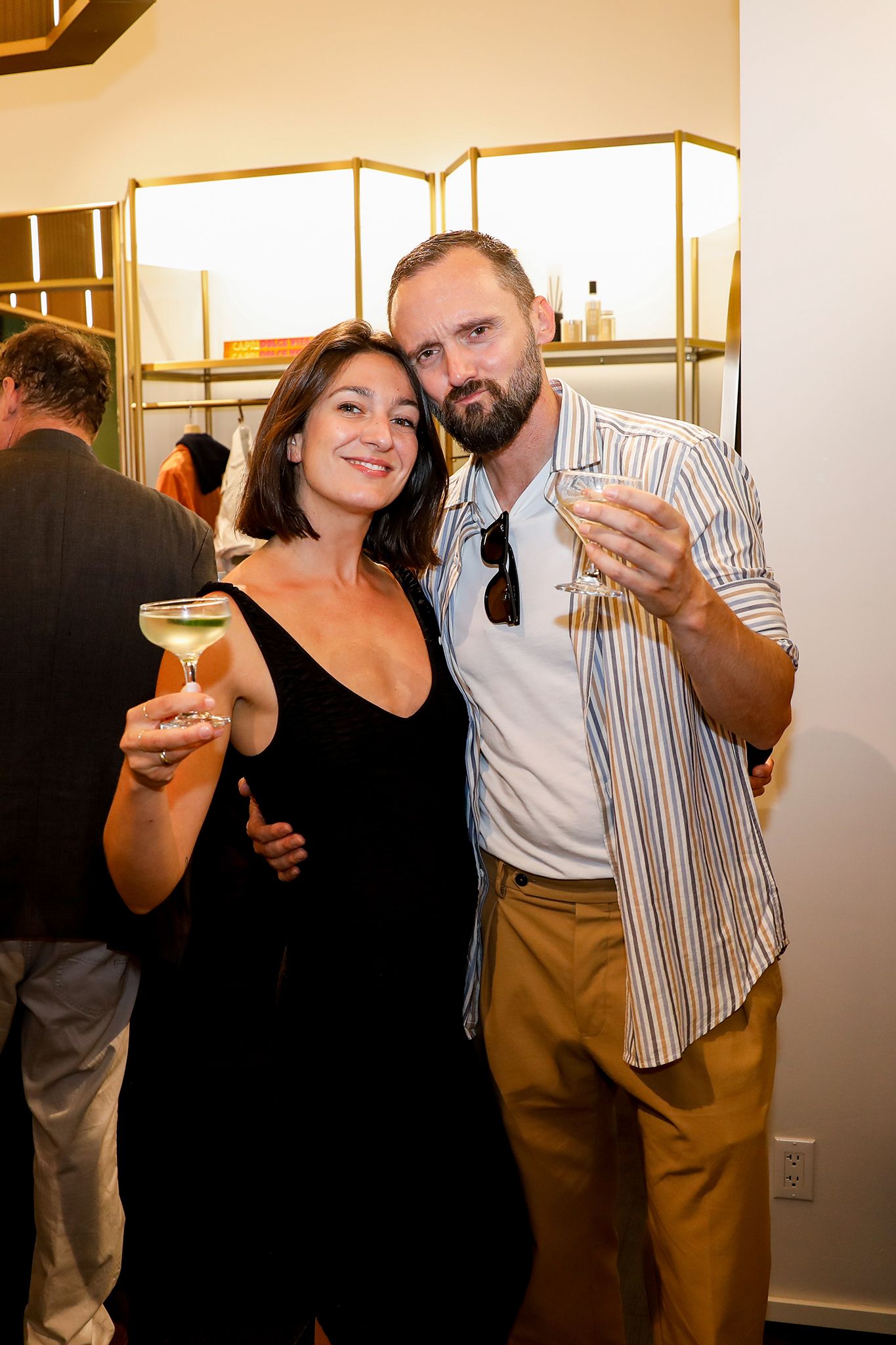A man and woman embrace, holding cocktails and looking at the camera.
