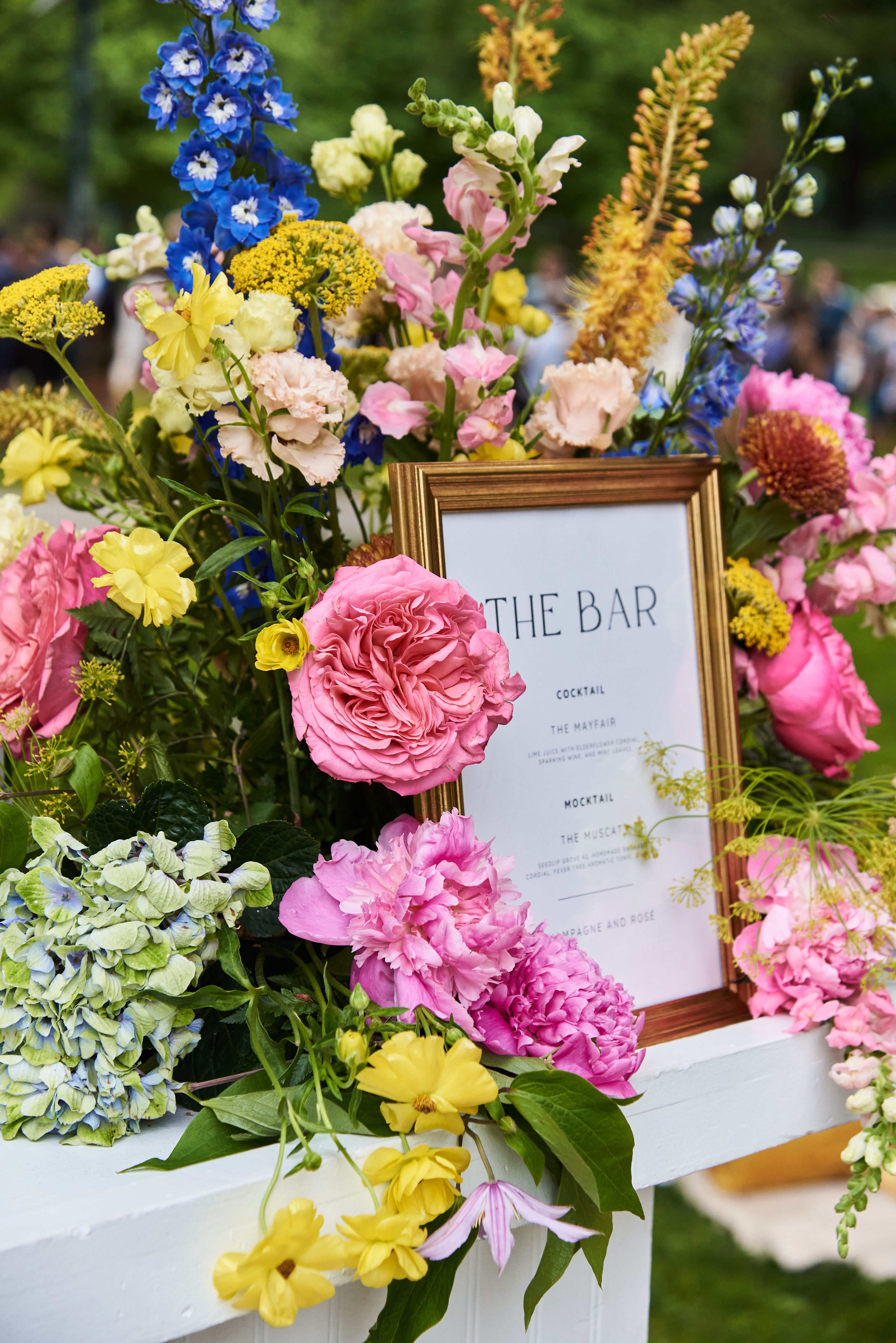 Floral bar display with cocktail menu sign at an outdoor catering event with colorful flower arrangements.