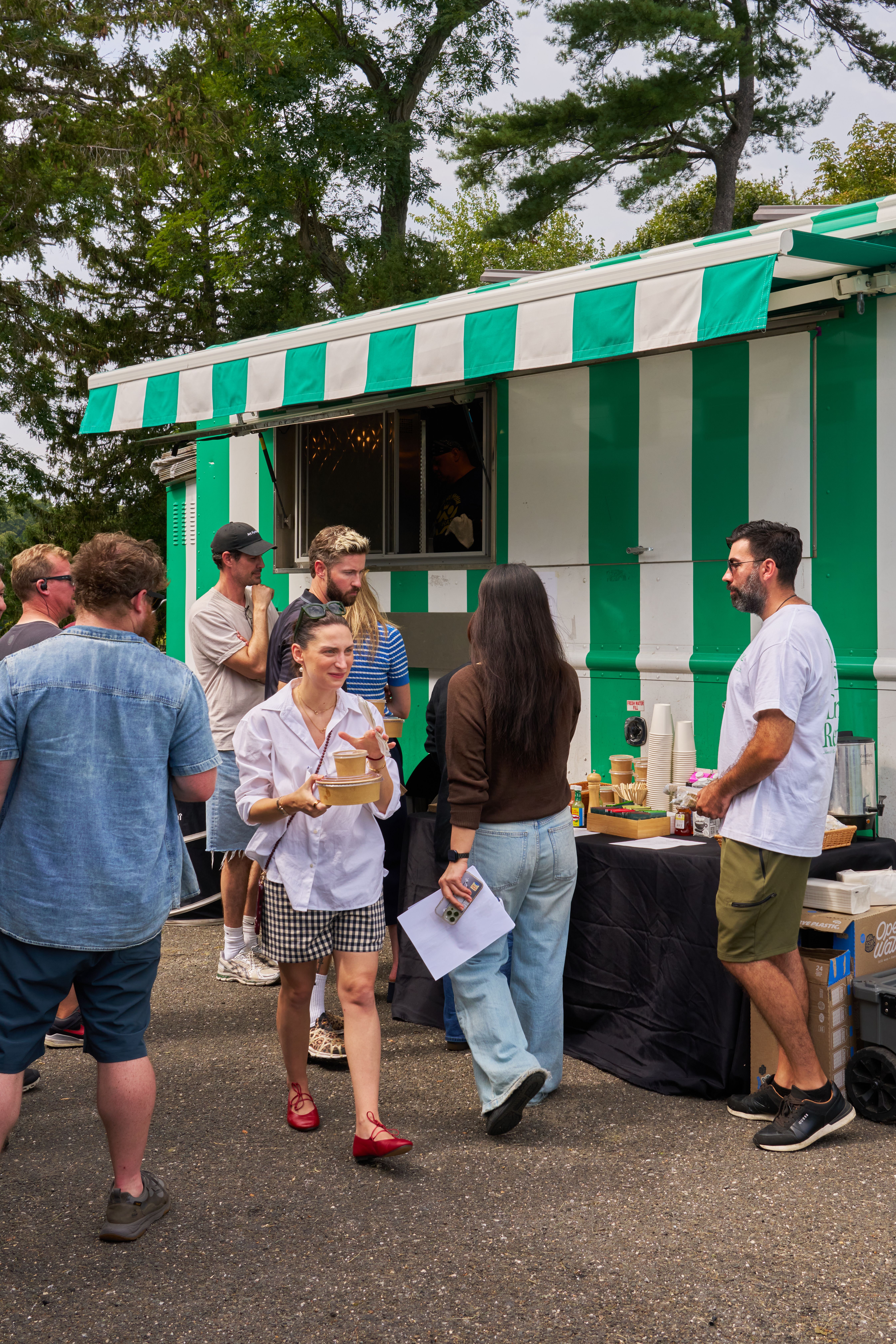 People gather around a green and white striped food truck; a woman in the foreground holds two meal containers.