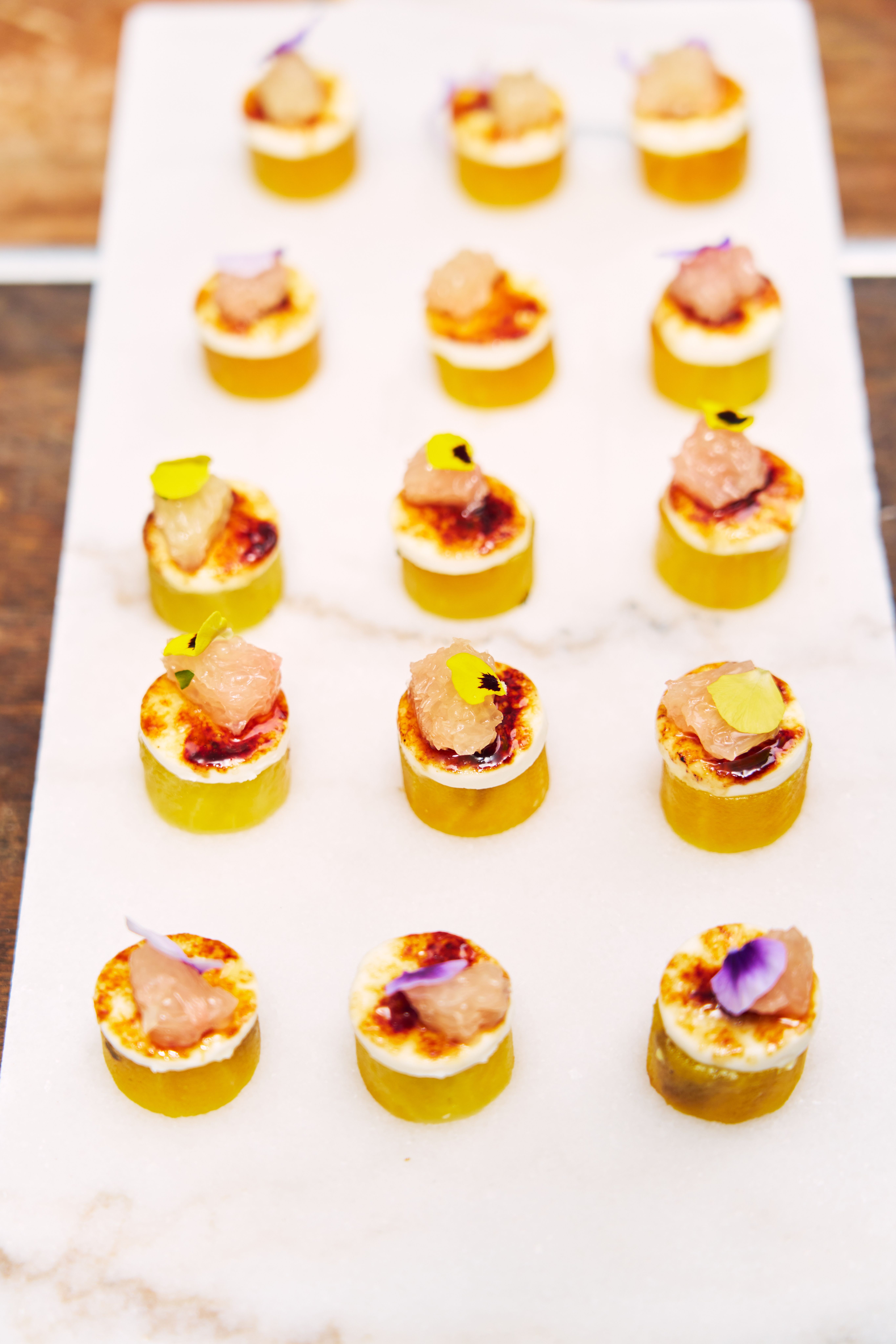 A wooden serving board with eight appetizers, each topped with cured meat, white cheese, and a basil leaf, held by two people.