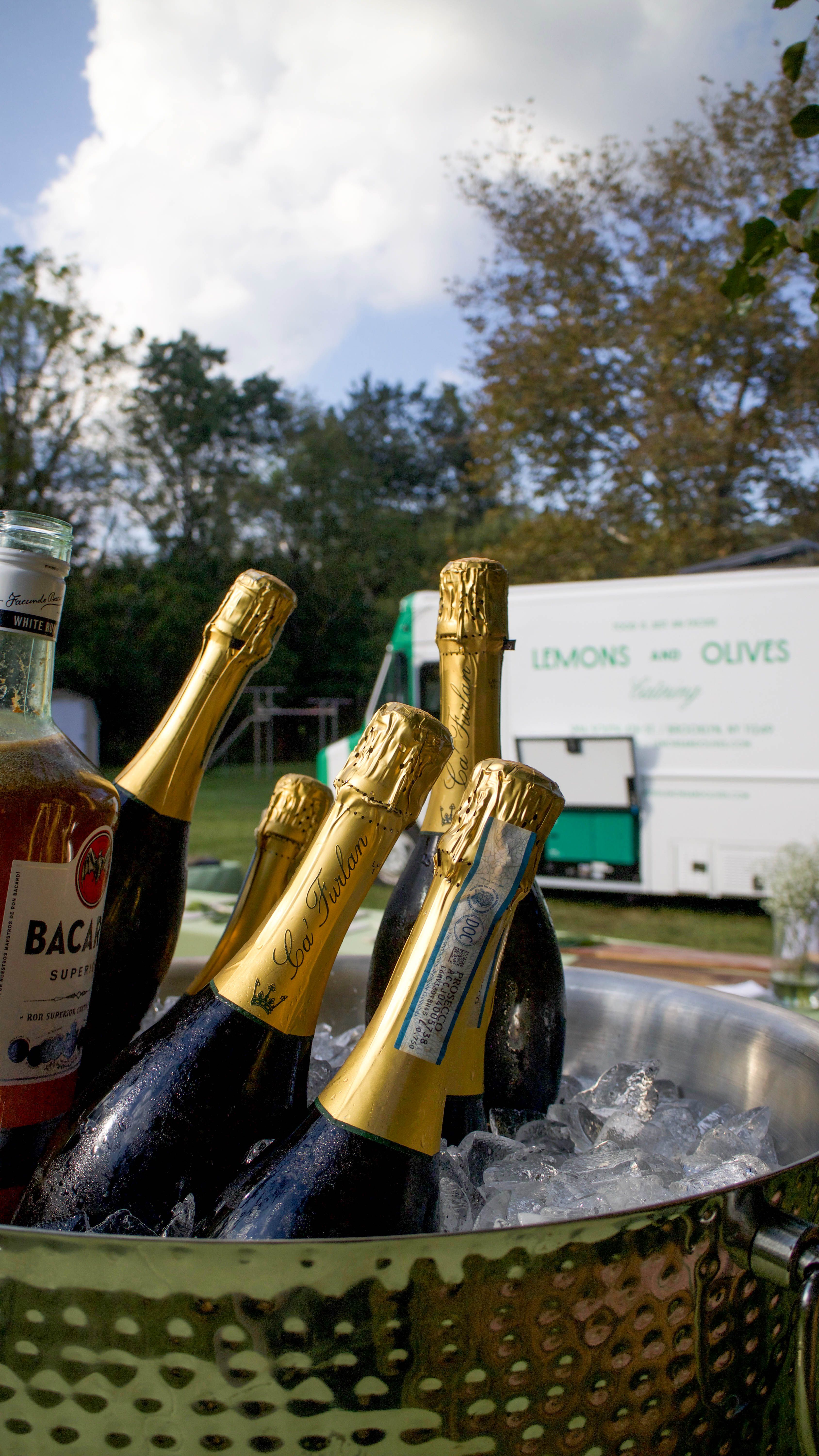 Bottles of sparkling wine and rum chill in a hammered metal ice bucket, with a catering truck and trees in the background.