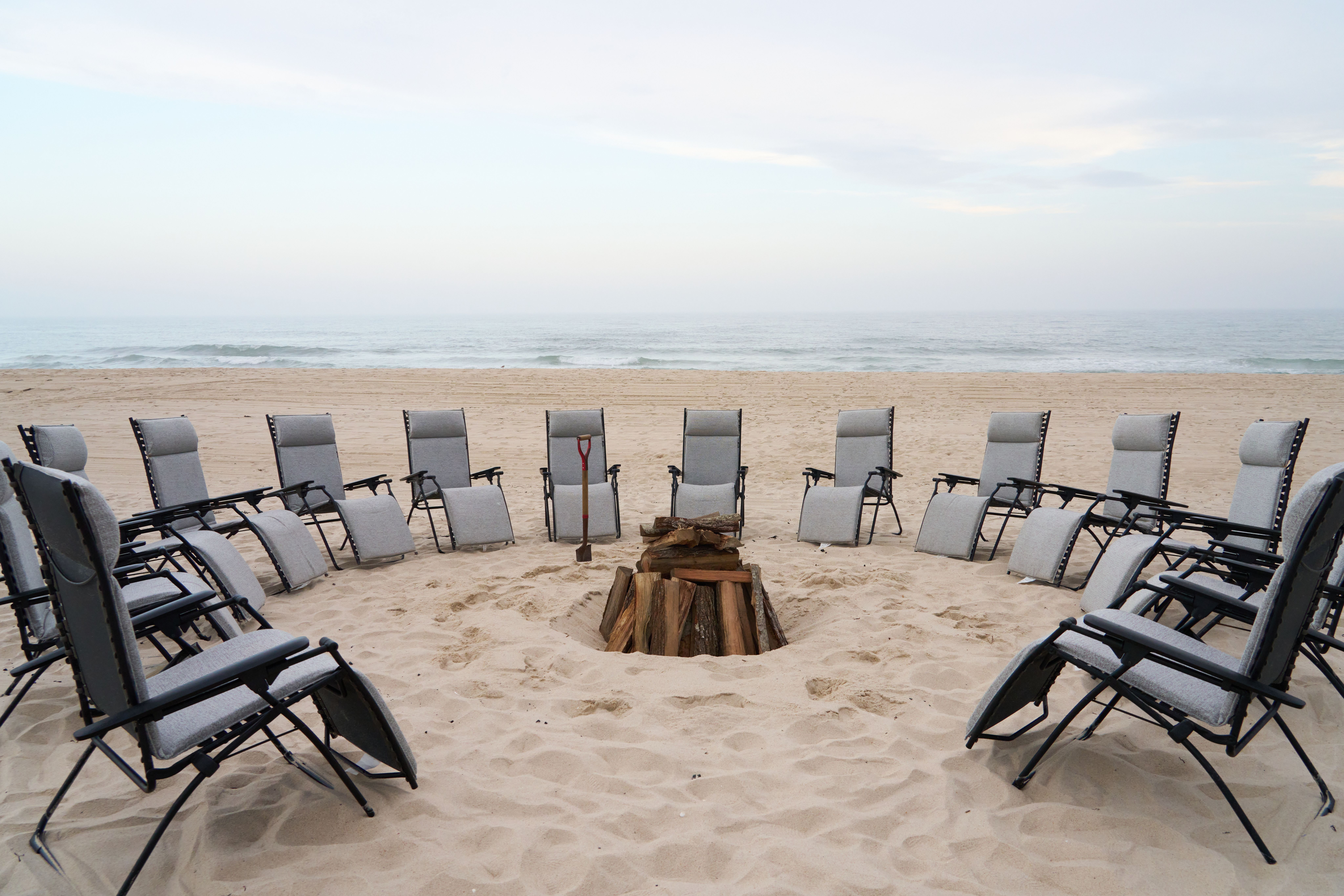 A circle of empty grey lounge chairs surrounds a stack of firewood and a shovel on a sandy beach with the ocean in the background.
