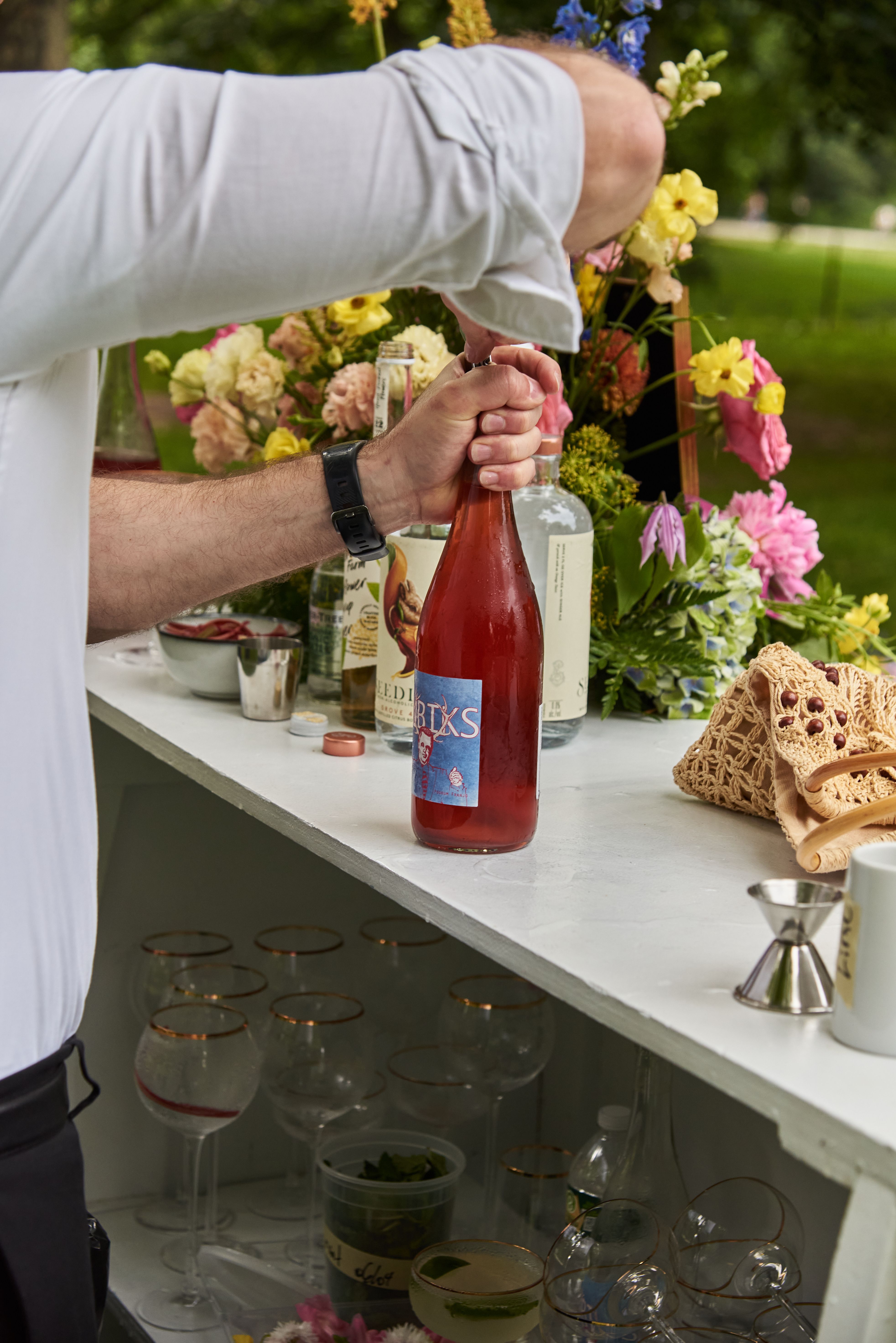 Bartender opening a bottle of rosé wine at an outdoor catered event bar with floral décor.