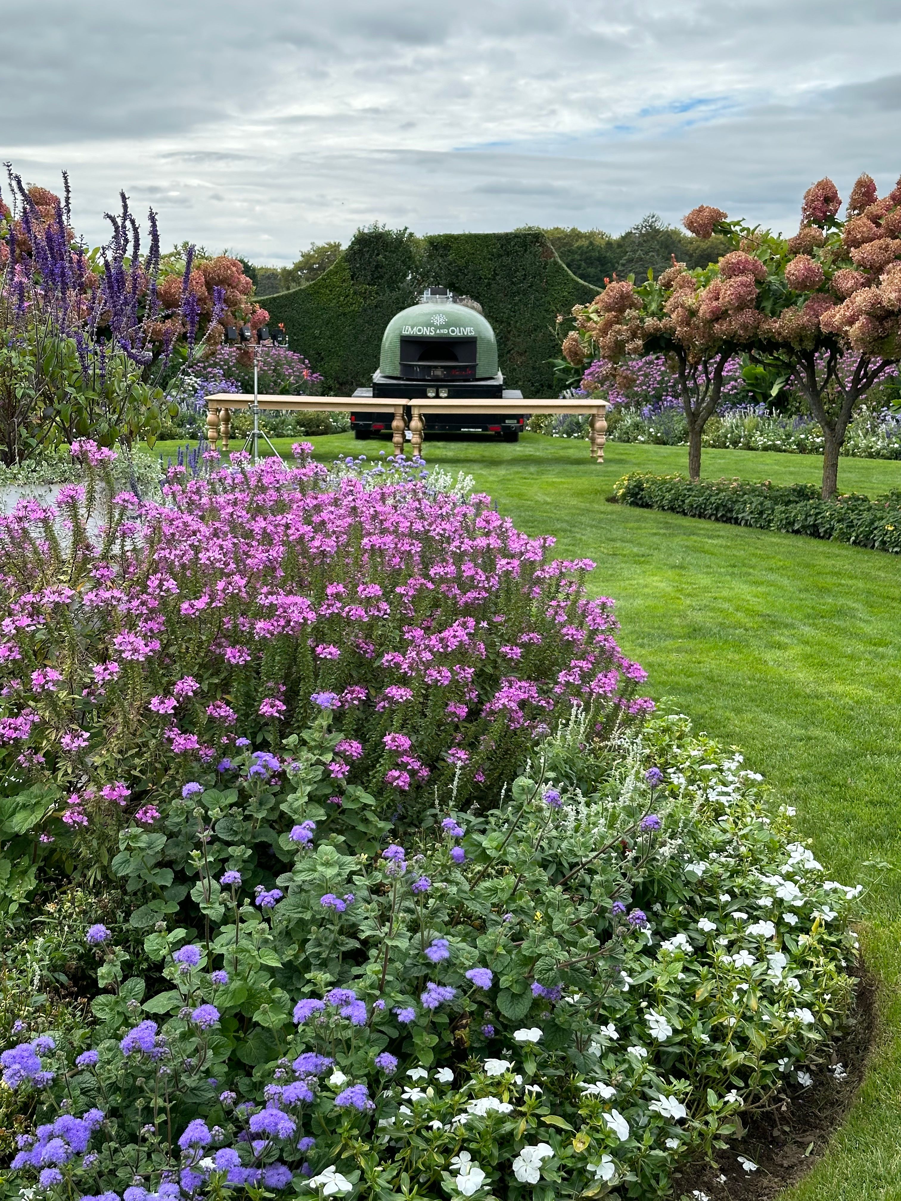 A dark green mobile pizza oven with a long wooden table sits in a lush garden filled with purple, pink, and white flowers.