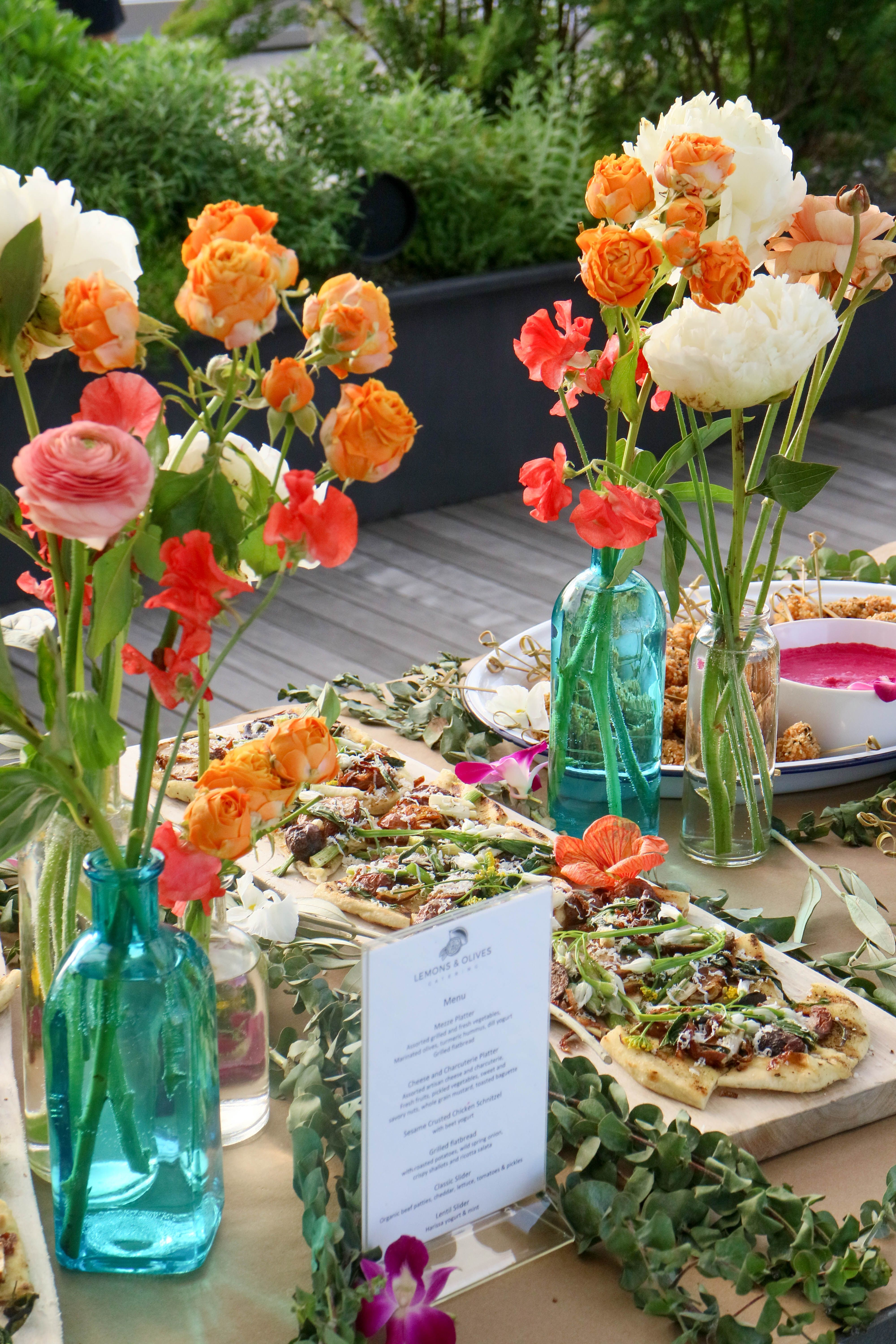 A table at an outdoor event, decorated with colorful flower bouquets, green garlands, flatbreads, and a "Lemons & Olives" menu.