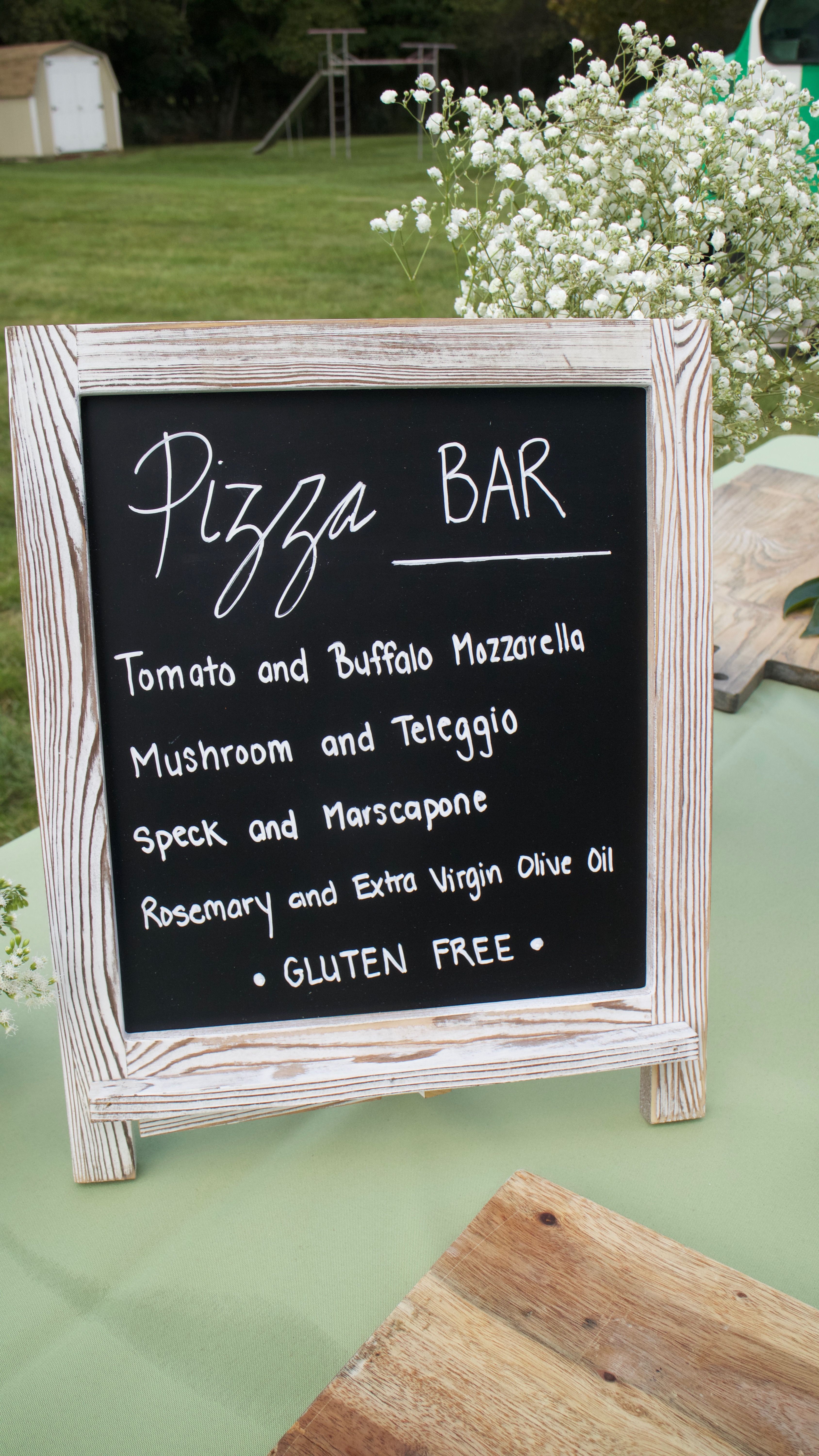 A chalkboard sign in a distressed white frame displaying a "Pizza BAR" menu with topping options and a "Gluten Free" note.