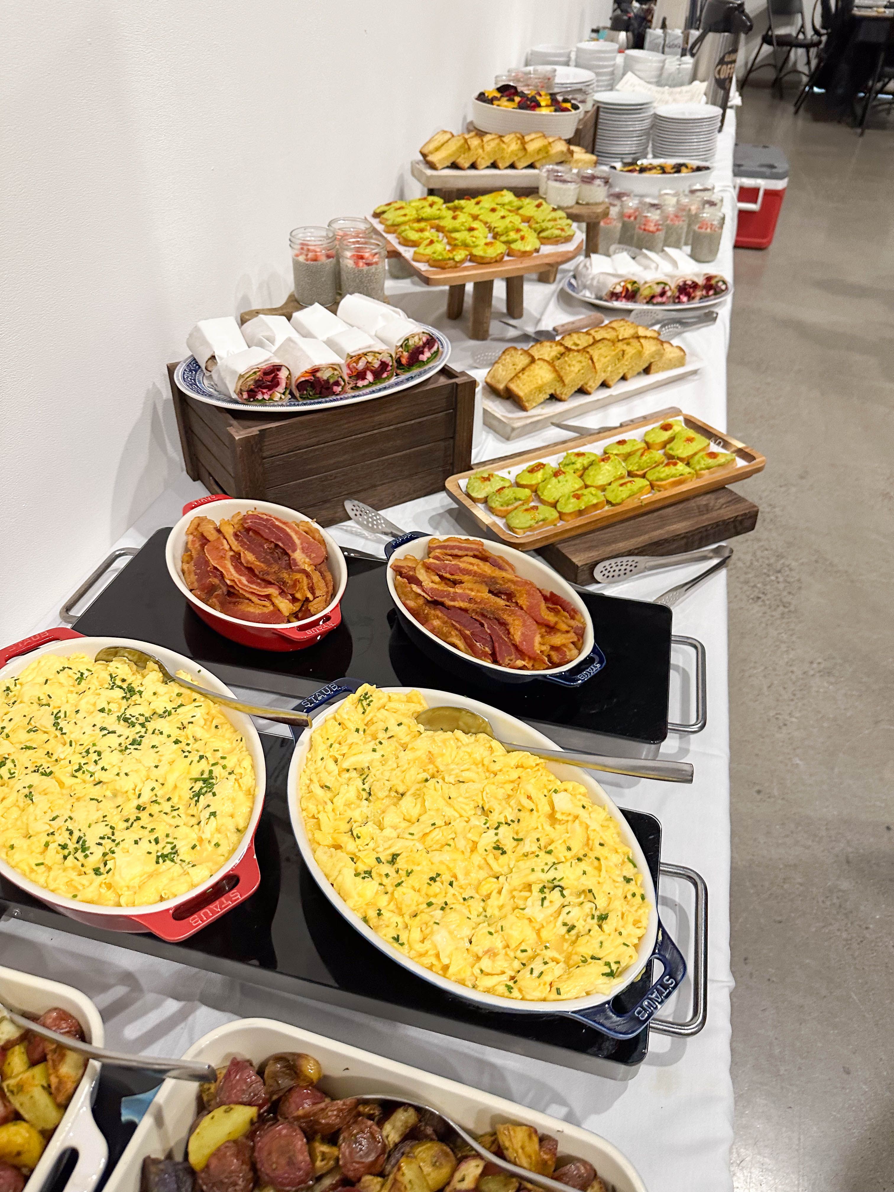 A long breakfast buffet featuring scrambled eggs, bacon, roasted potatoes, wraps, pastries, fruit, and individual parfaits.