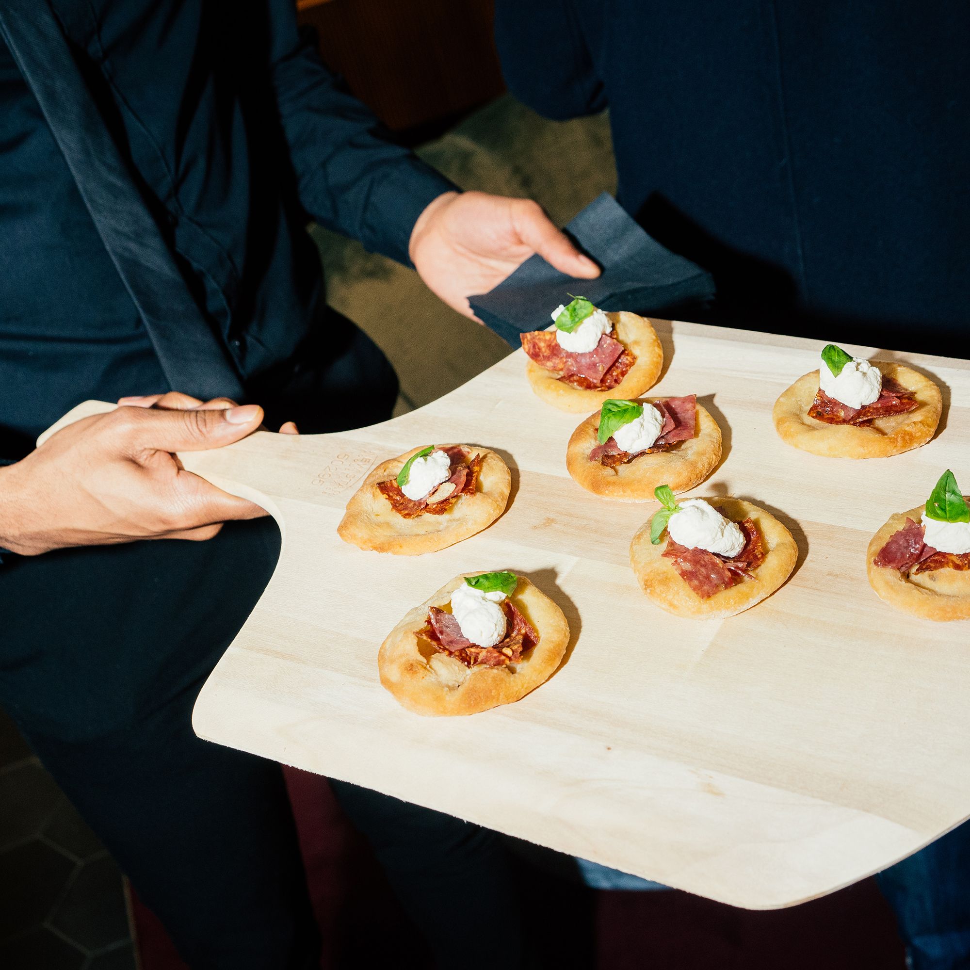 Catering staff serving passed flatbread canapes and hors d'oeuvres topped with cured meat and ricotta during cocktail hour.