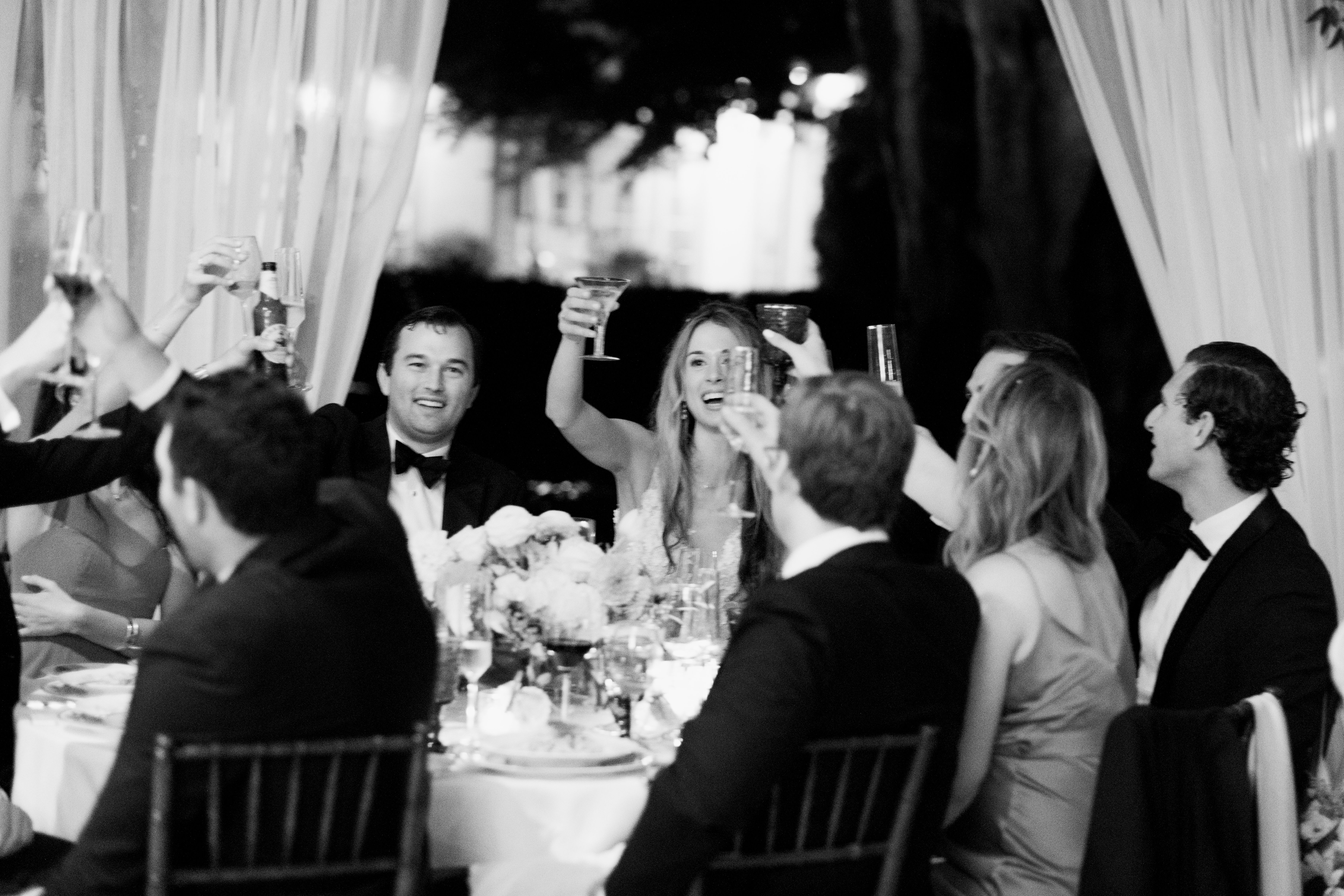 Black and white photo of formally dressed guests raising their glasses for a toast at a table.